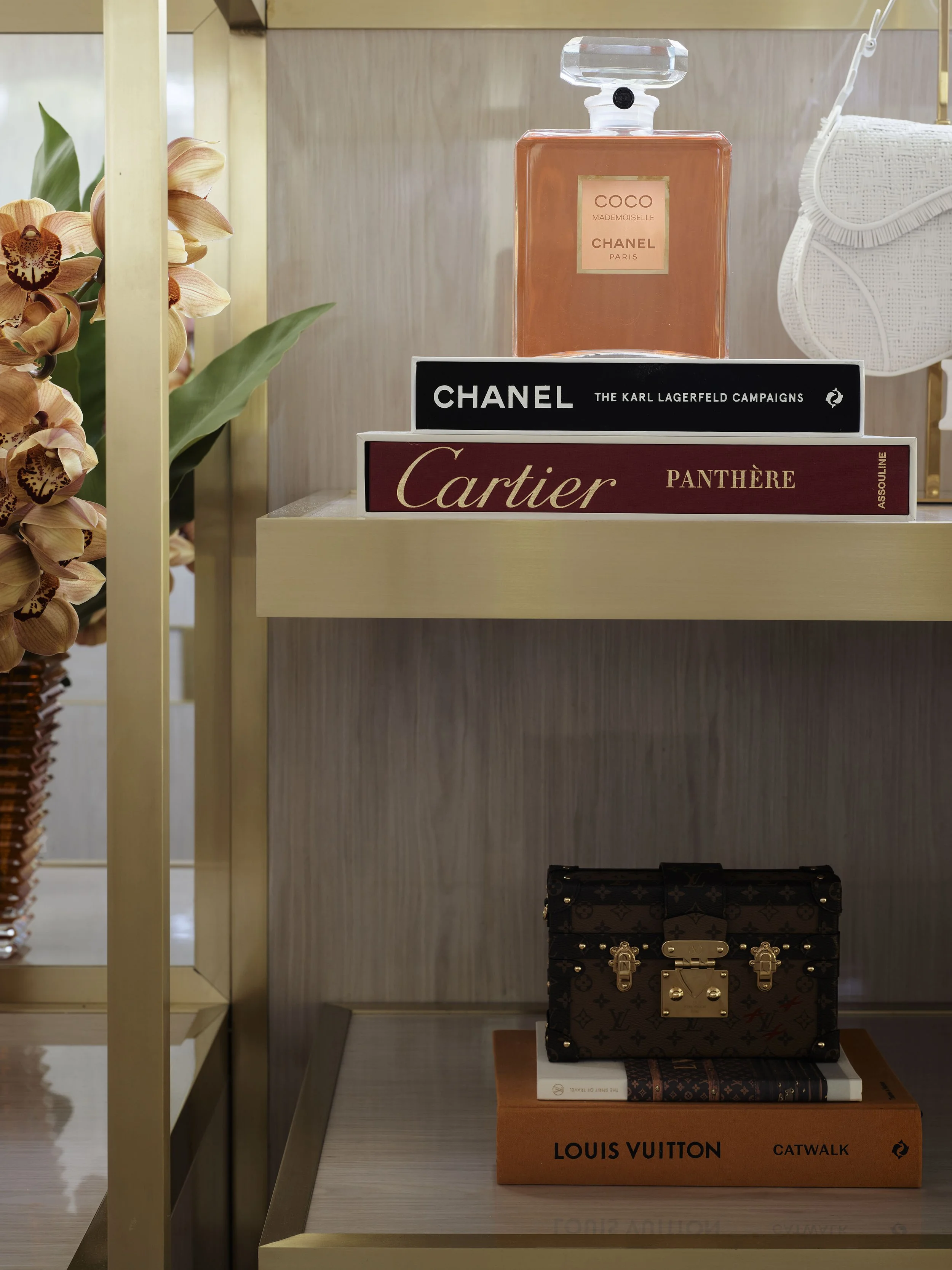 A shelf displaying a Louis Vuitton handbag on a Louis Vuitton book, two other fashion books, a large Chanel book, a Cartier book, and a Chanel perfume bottle on top of the books.