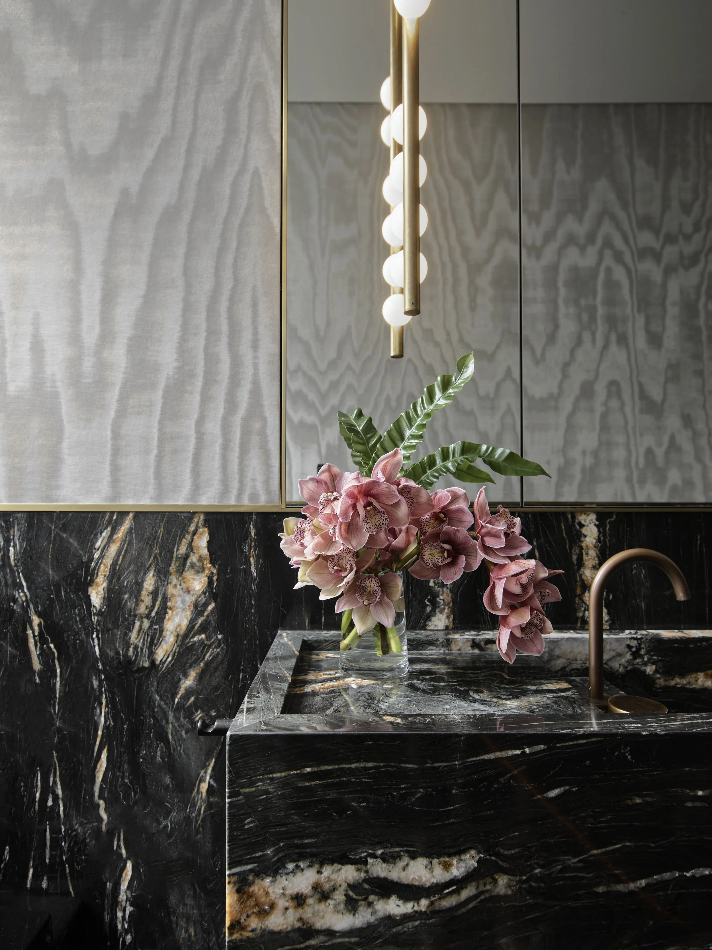 A bathroom countertop made of black marble with white and gold veining, a glass vase with pink orchids and green leaves, a gold faucet, and a large mirror with modern light fixtures above.