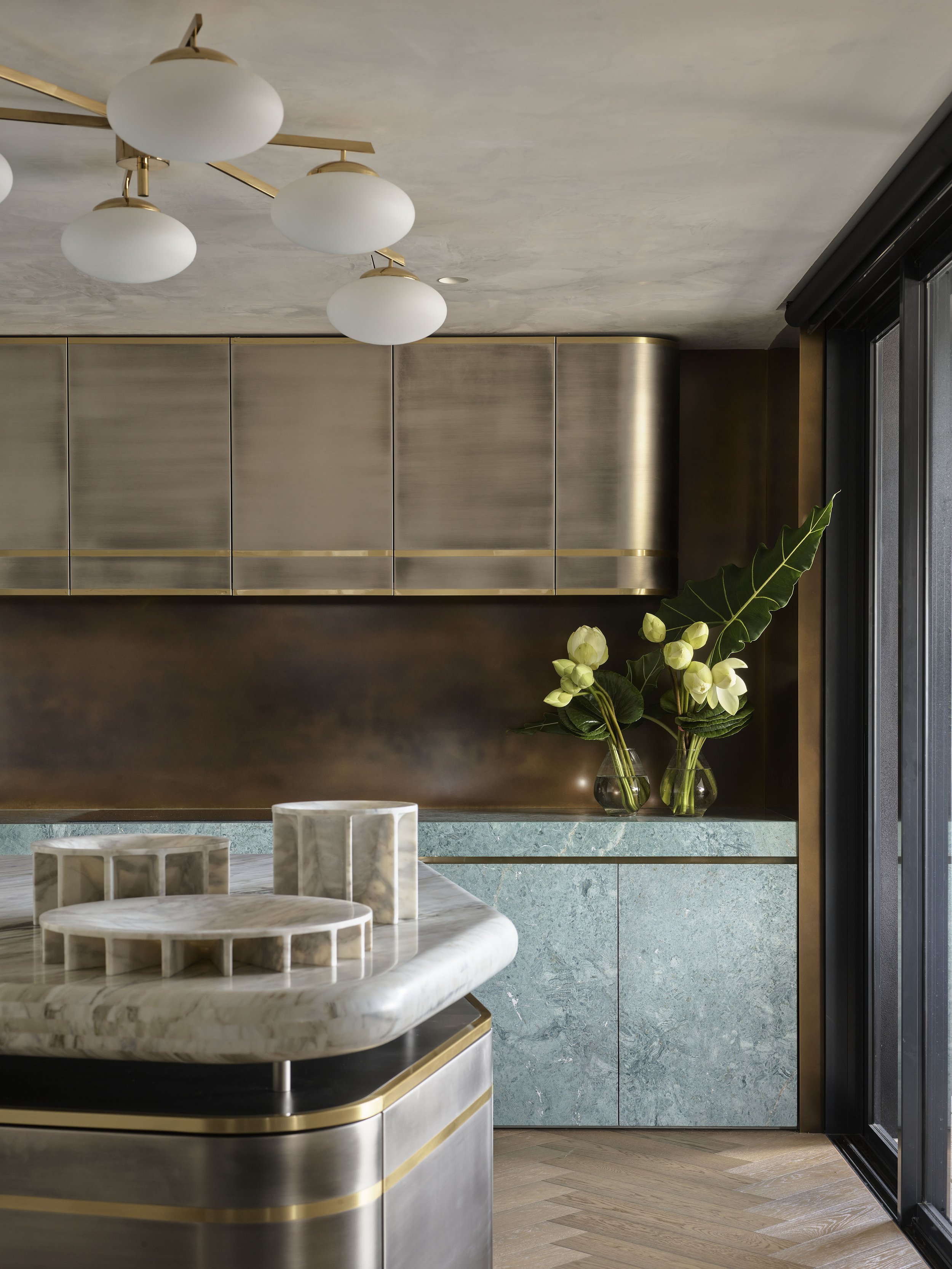 Modern kitchen with gold and stainless steel cabinets, green marble countertops, a marble island with geometric decor, and a large window with black framing, decorated with white flowers and large green leaves.