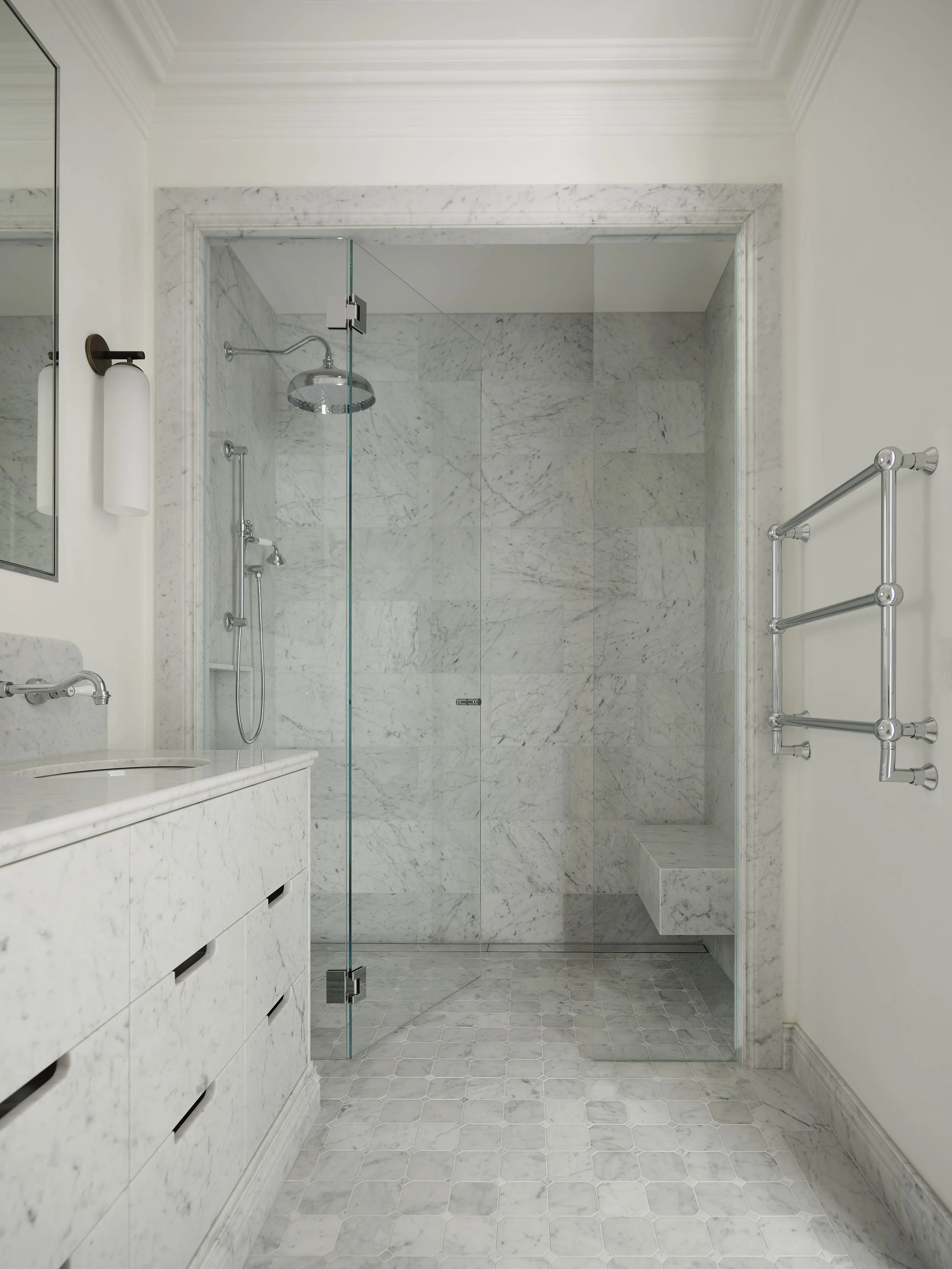 A bathroom with a marble shower enclosure, glass door, a rain shower head, a handheld shower, and built-in marble bench. White marble vanity with a mirror and a wall-mounted light fixture.