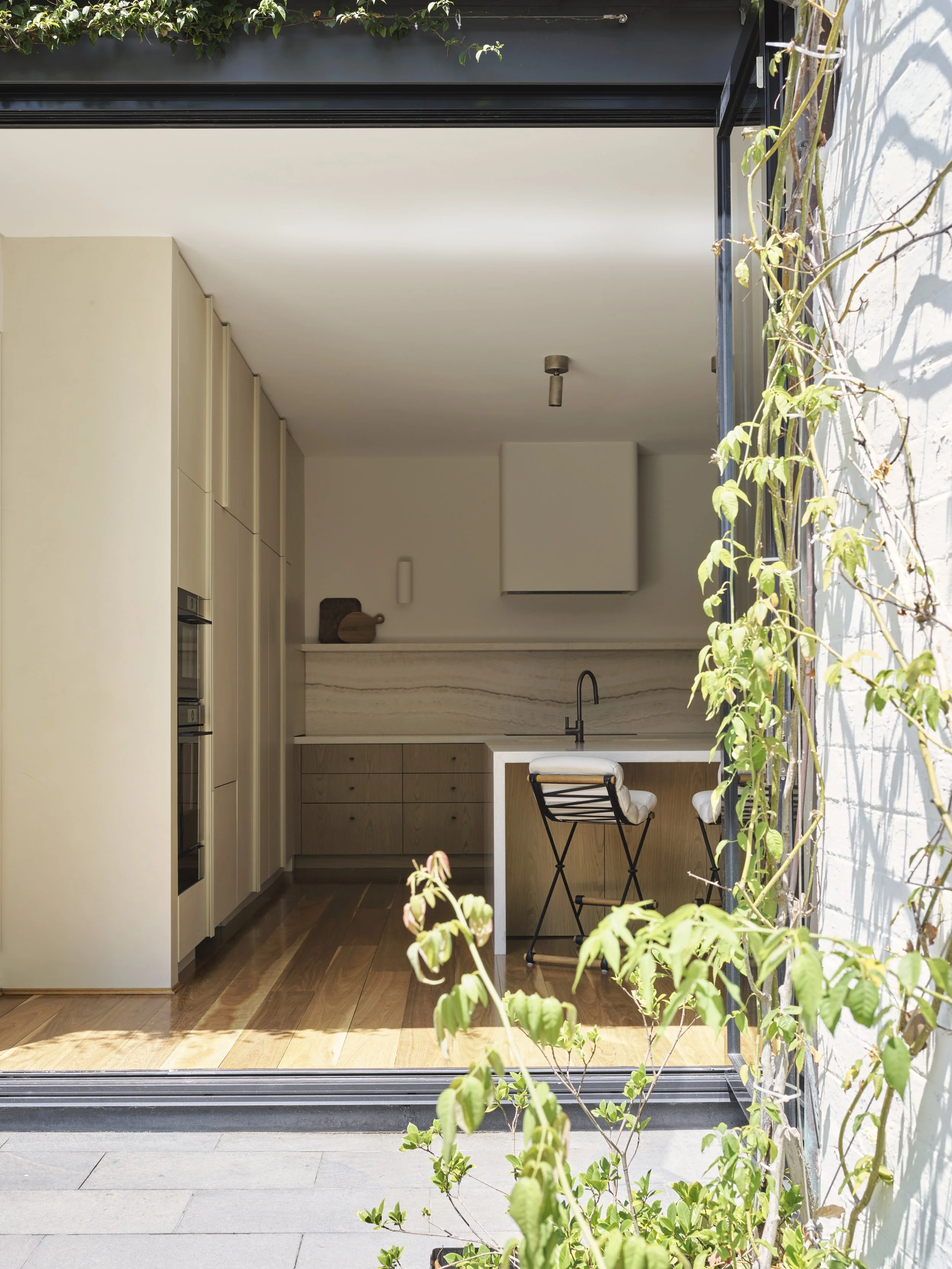 View of a modern kitchen seen through an open sliding glass door, featuring wooden cabinetry, a white countertop, a black faucet, two black chairs with white cushions, and green plants creeping along the right side.