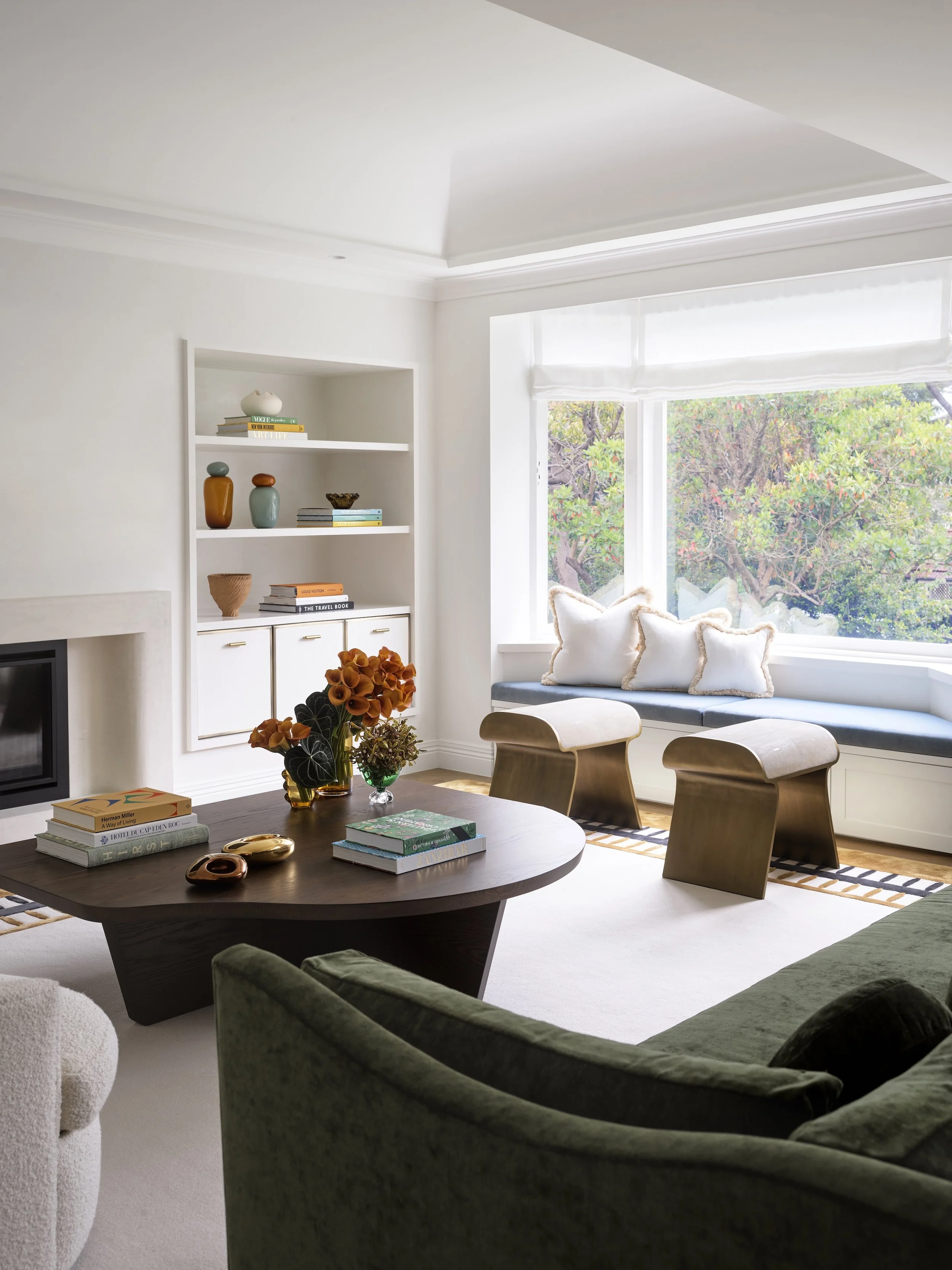Bright living room with large window, built-in window seat with pillows, wooden coffee table with books and flower arrangements, green armchair, and white built-in shelves with decorative items.