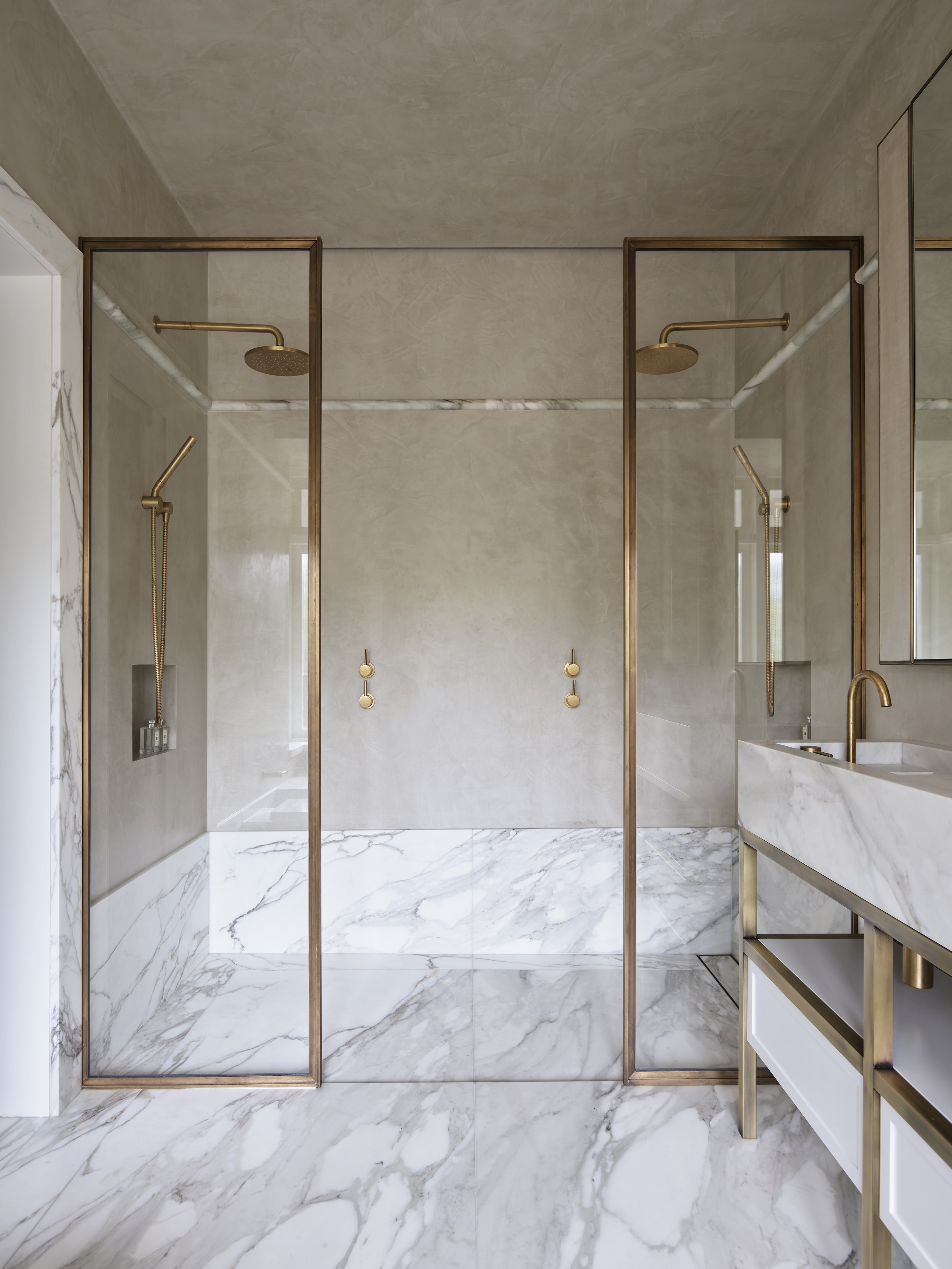 Luxury bathroom with two glass-enclosed walk-in showers featuring gold fixtures and marble accents, and a marble vanity with a gold faucet.