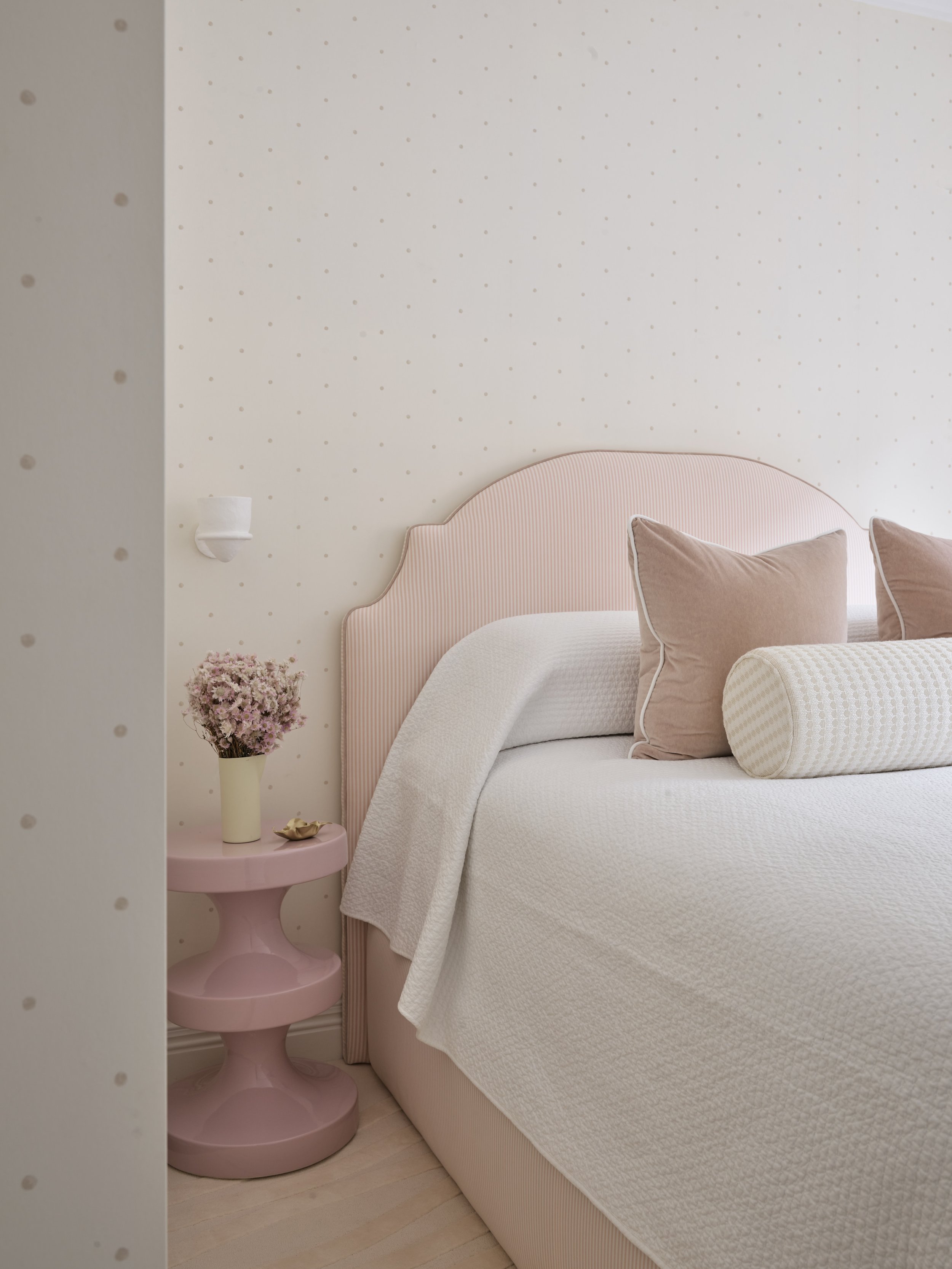 A cozy bedroom scene with a pink upholstered headboard, beige pillows, a white quilt, and a pink side table with a vase of pink flowers.