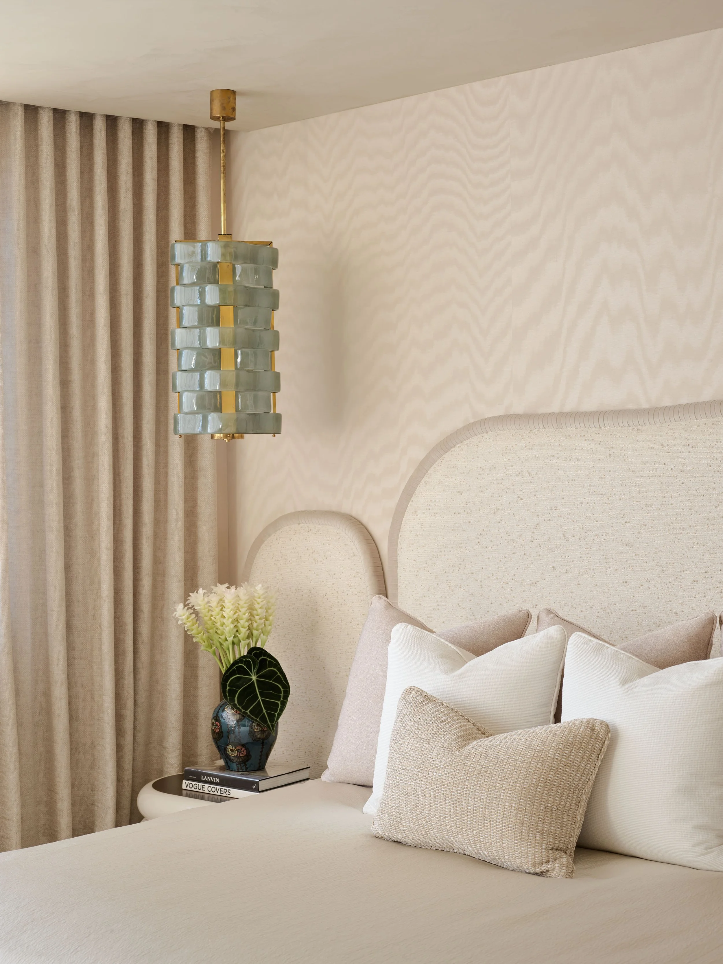 A bedroom with a beige bed, multiple pillows, a side table with a flower vase, and a hanging pendant light.