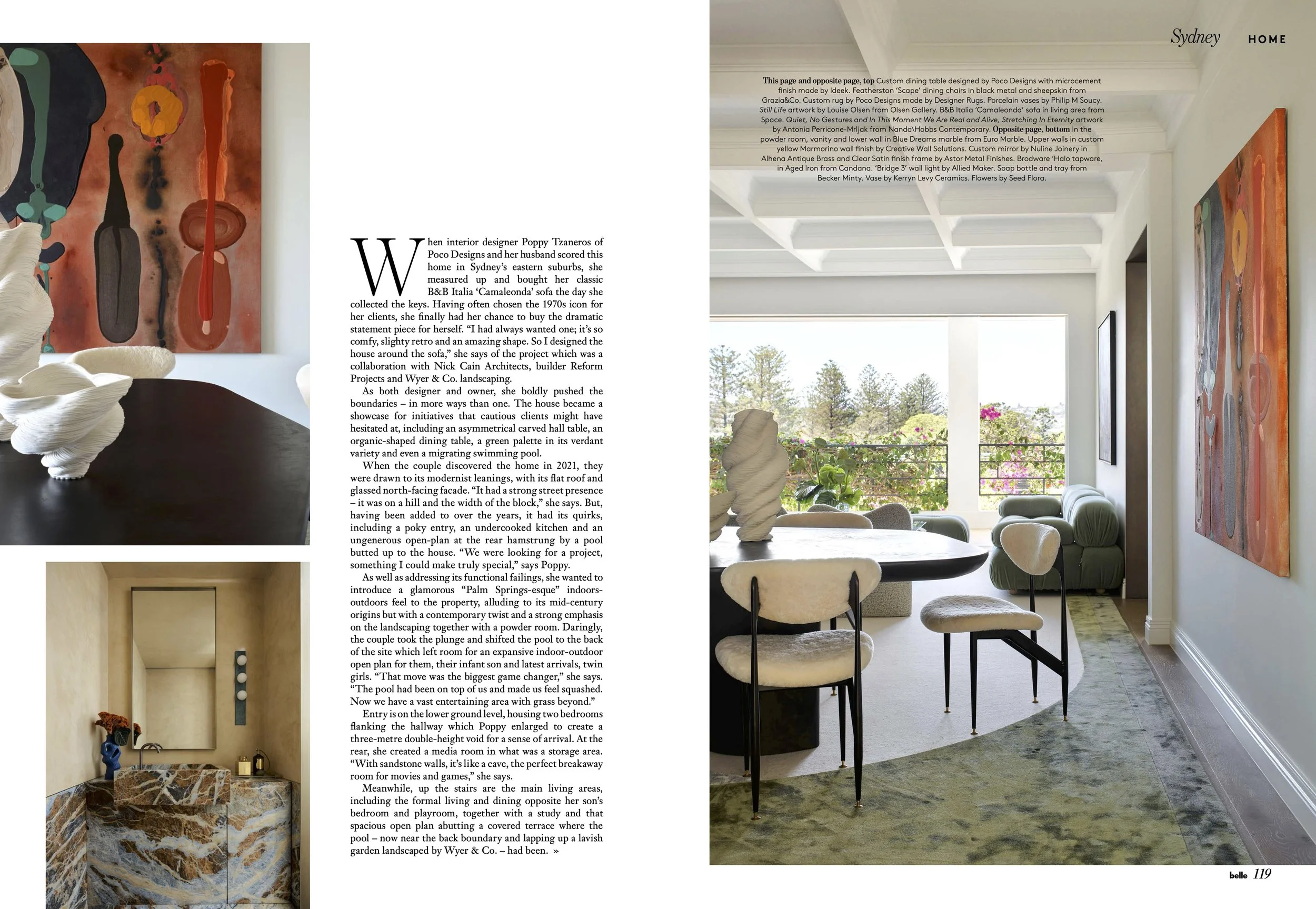 A spacious, modern living room with large windows overlooking an outdoor balcony garden. The room features contemporary furniture, abstract artwork on the walls, and decorative sculptures on a table. There is a mix of light and dark tones, with a green rug, white beams on the ceiling, and outdoor greenery visible through the windows.