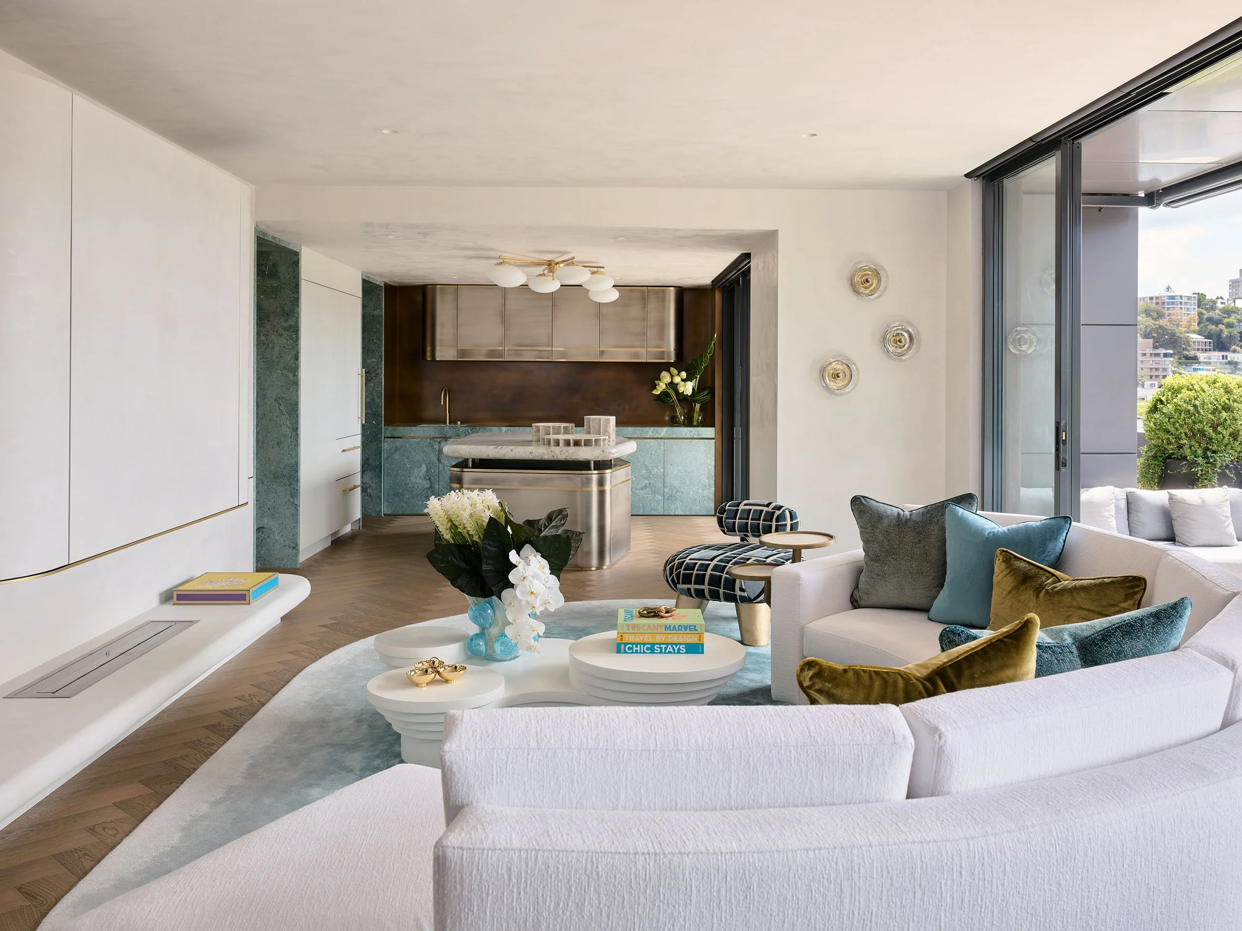 Modern living room with a white curved sofa, colorful pillows, a patterned chair, a white coffee table with books and decorations, and large sliding glass doors opening to a balcony with city views.