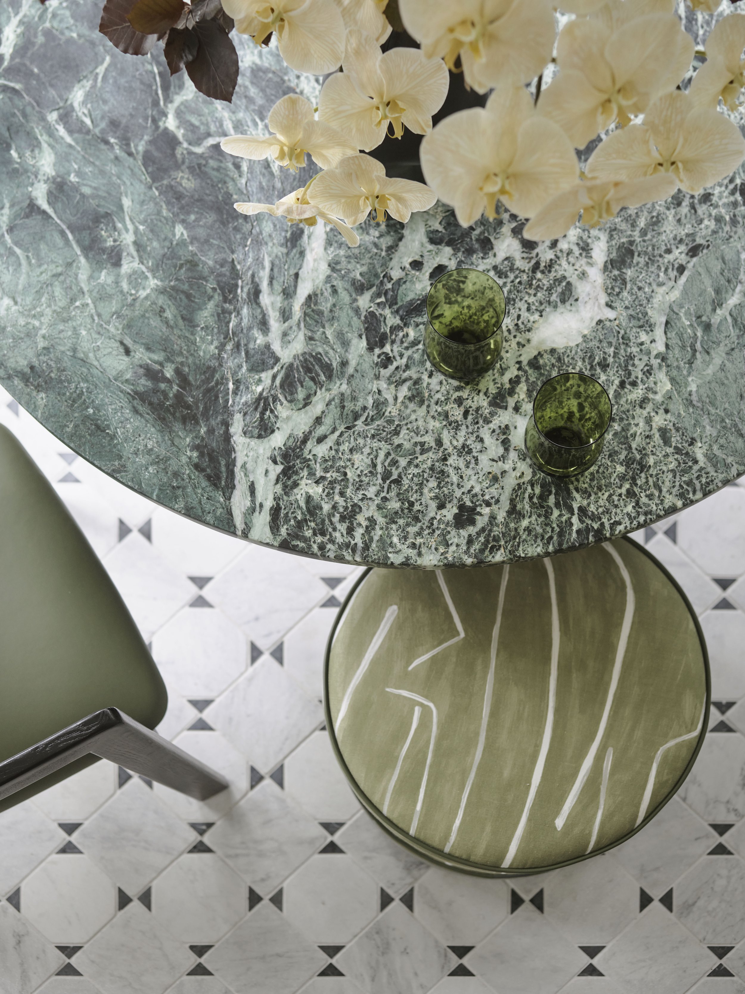 Top-down view of a round green marble table with a floral arrangement of white orchids and two green glasses, positioned above a patterned tile floor.