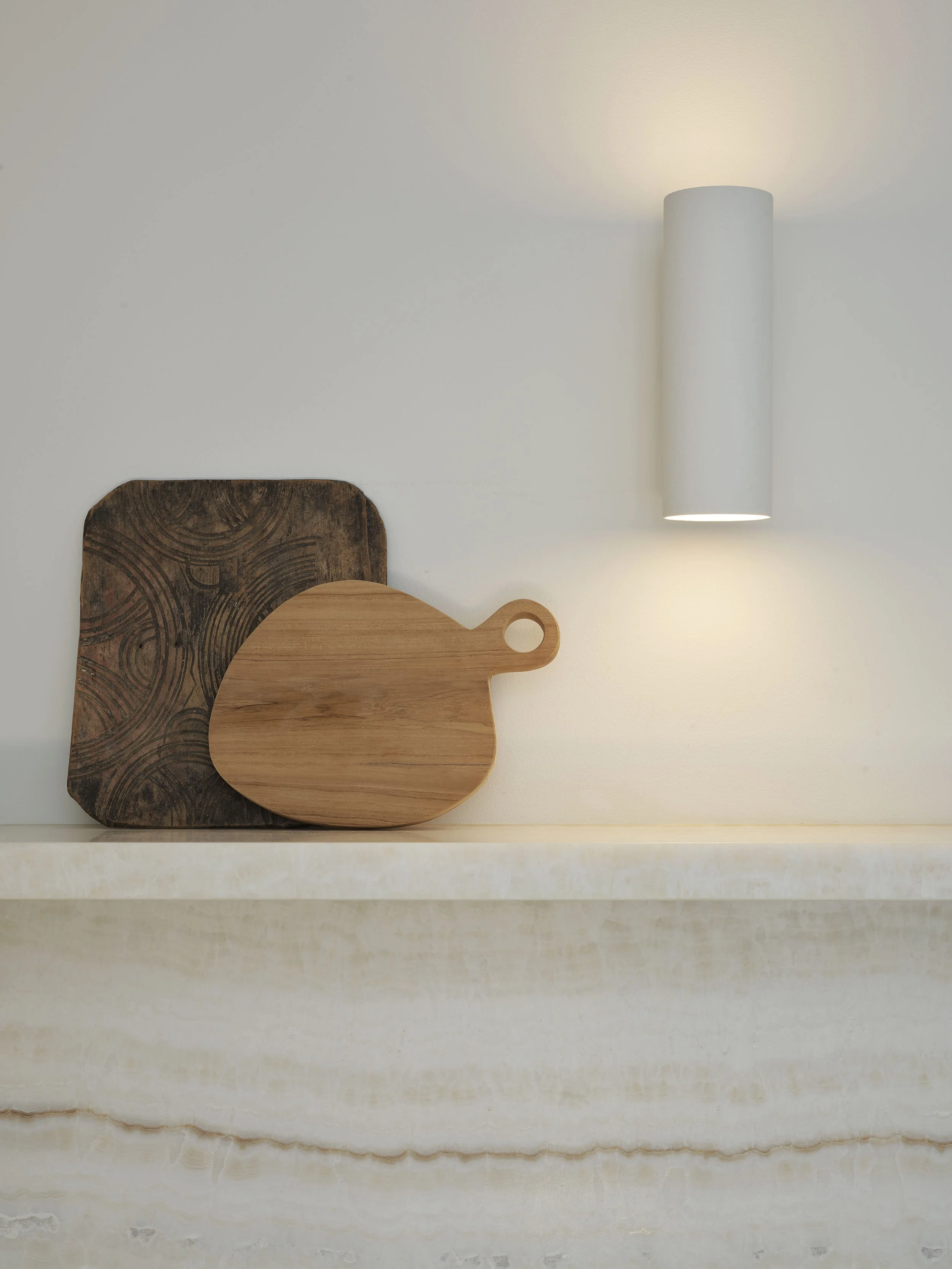 A white wall with a cylindrical wall lamp, a wooden cutting board, and a dark textured chopping board on a light-colored shelf.
