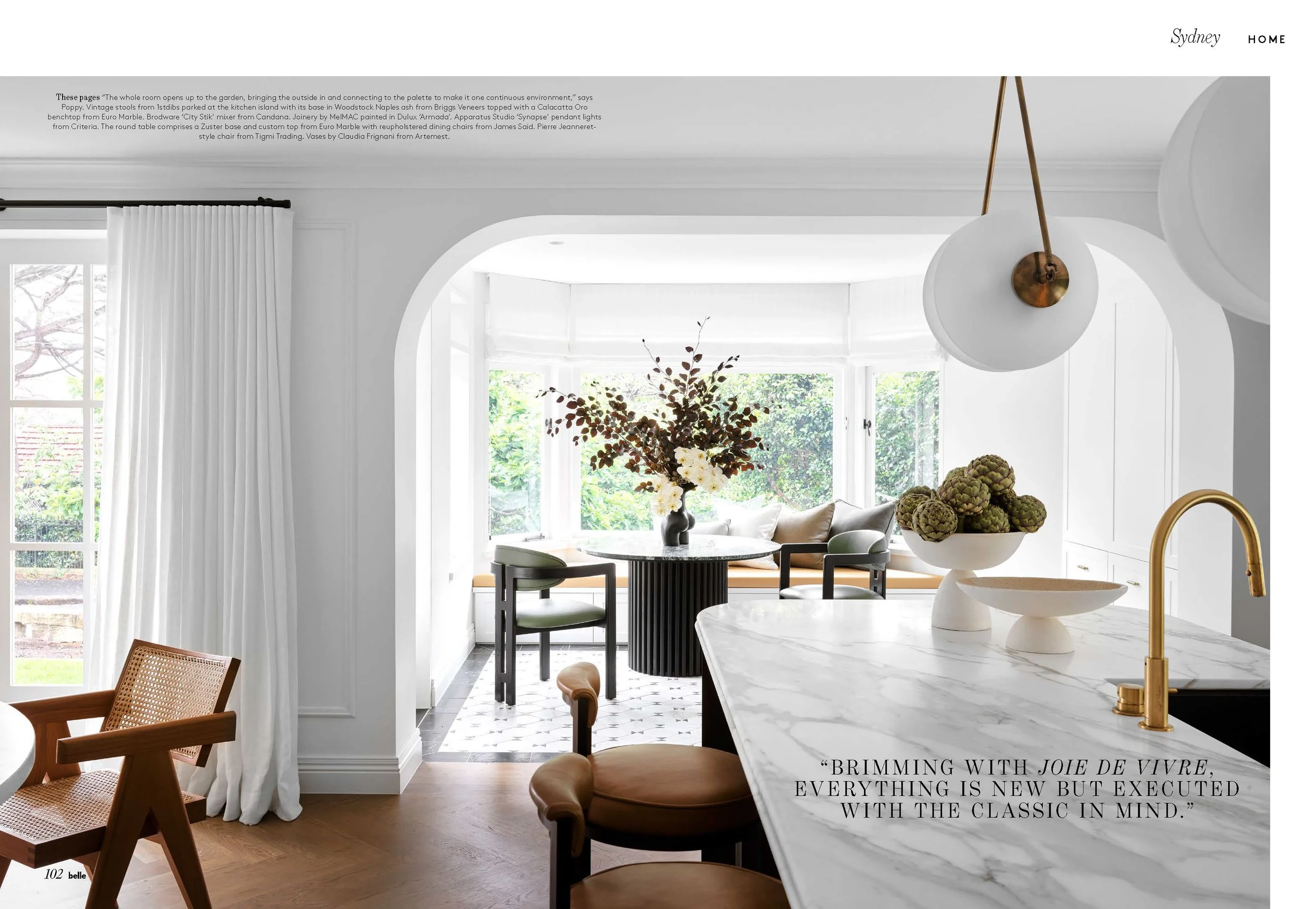 Bright interior view of a kitchen and dining area with white walls, large windows, and elegant furniture, including marble countertops, chairs, and decorative vases with flowers and artichokes.