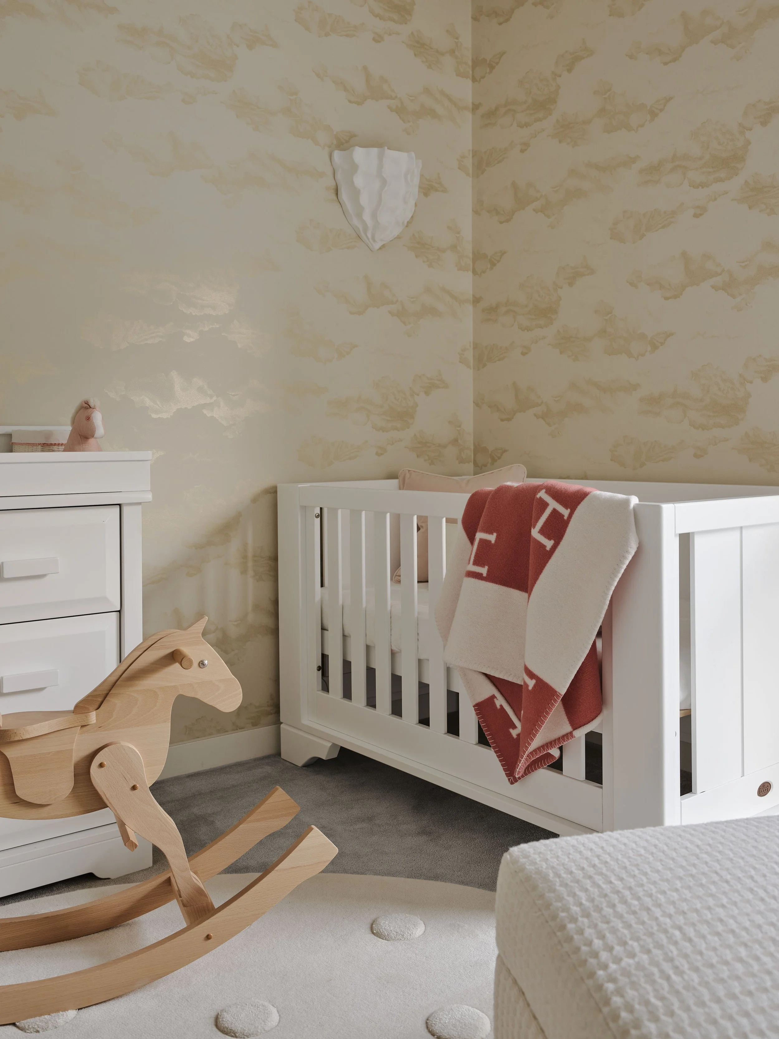 A nursery with a white crib, a wooden rocking horse, a white dresser, and a plush at the foot of the bed. Pink and white towels hang on the crib, and a decorative wall piece hangs above.