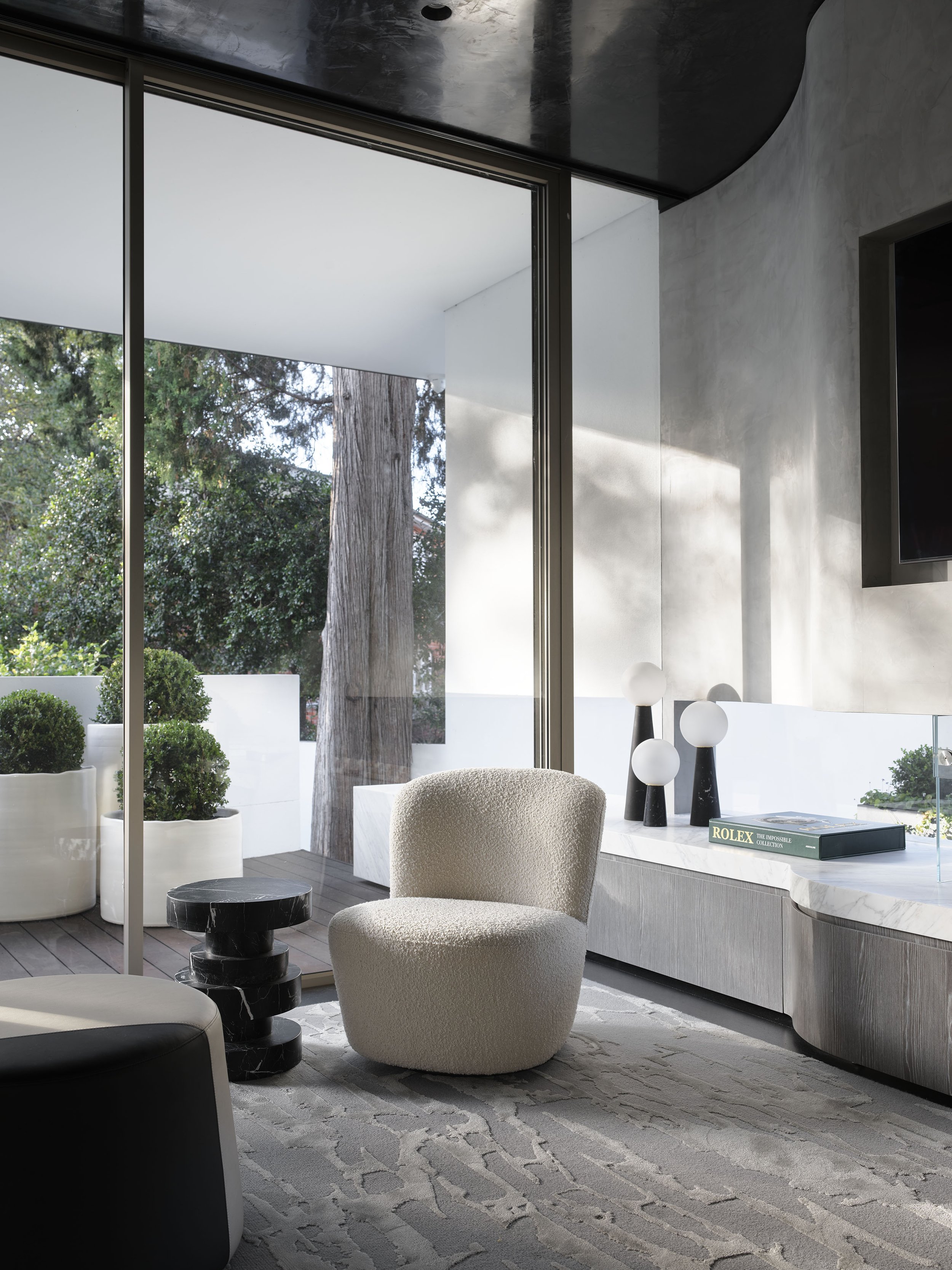 Modern living room with large glass sliding doors, beige textured armchair, black marble side table, abstract patterned gray rug, and minimalist decor including three black and white lamps and a book titled 'ROLEX' on a white marble console.
