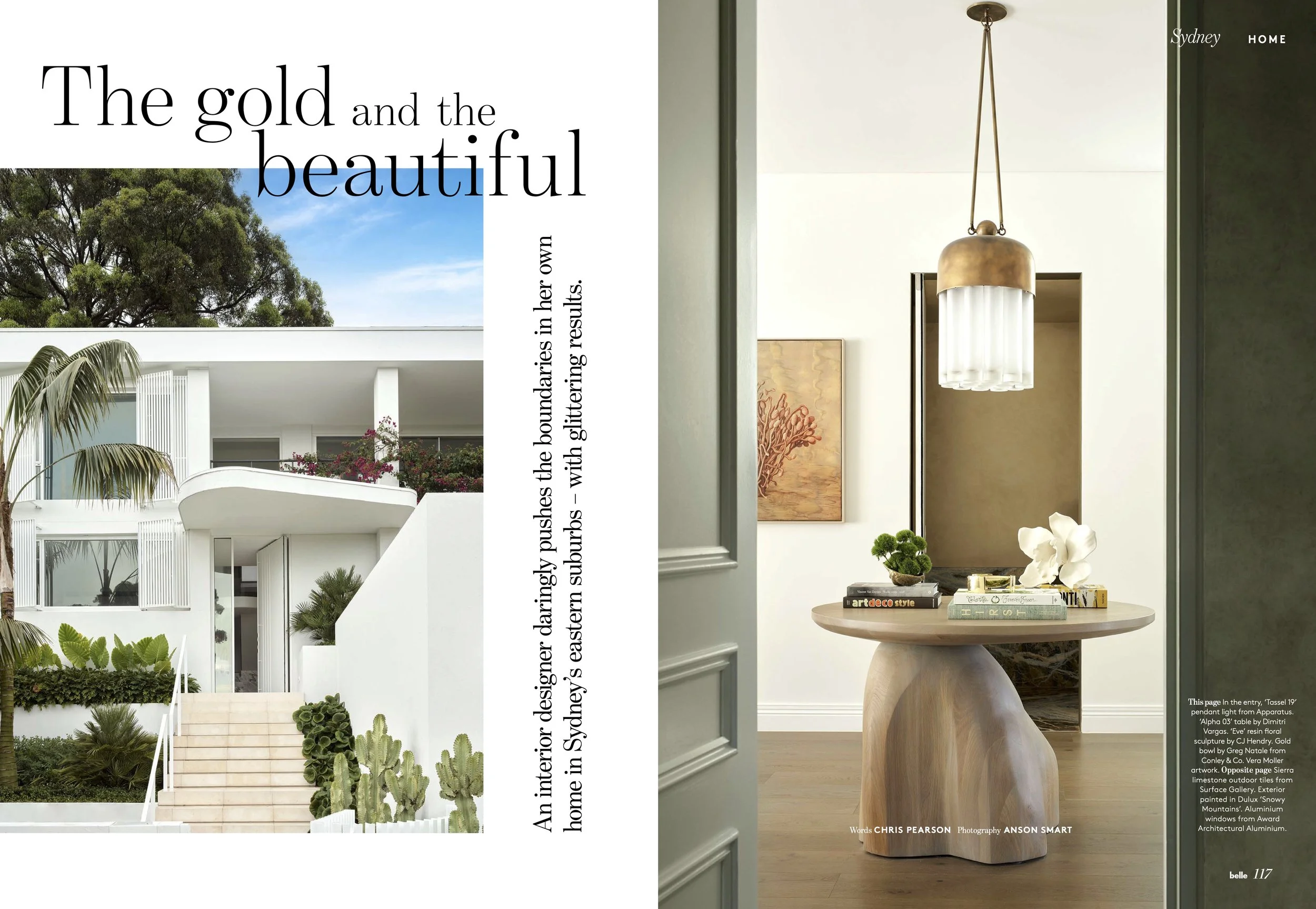 The left side shows the exterior of a modern white house with stairs, lush green plants, palm trees, and pink flowers, under a blue sky with clouds. The right side features the interior of a stylish room with a round wooden table decorated with a small bonsai tree, white flowers, and books, a large rectangular mirror, a hanging gold and glass pendant light, and a painting of red coral on the wall.