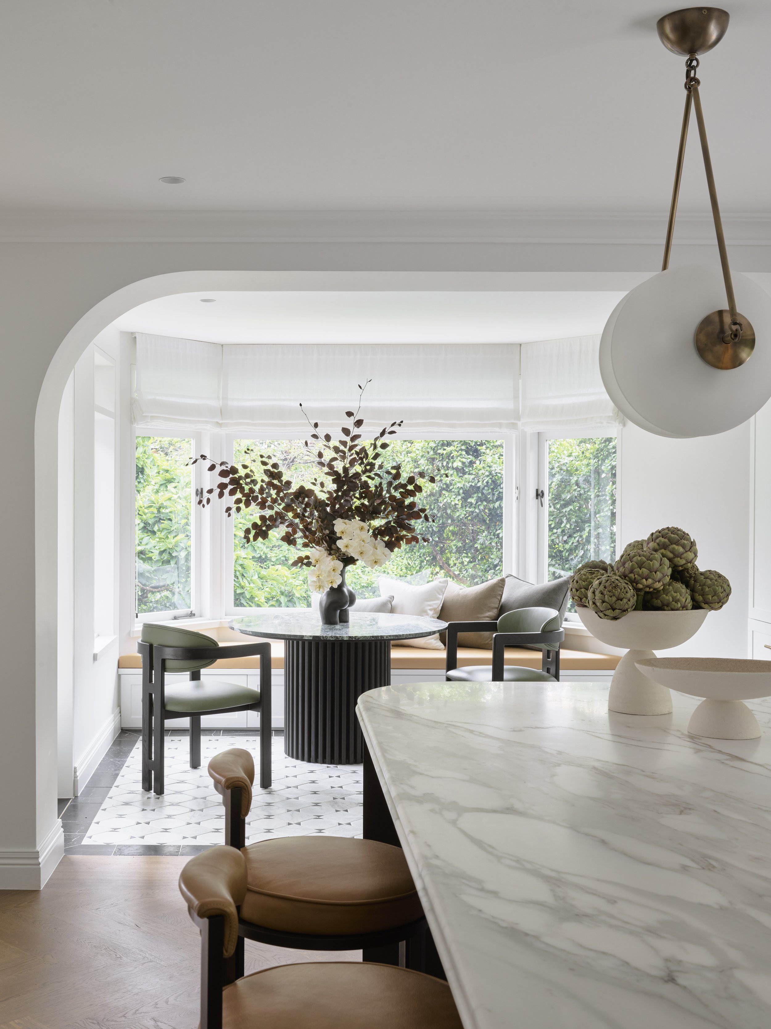 Bright dining nook with large bay window, cushioned seating, a round table with floral arrangement, and modern chairs.