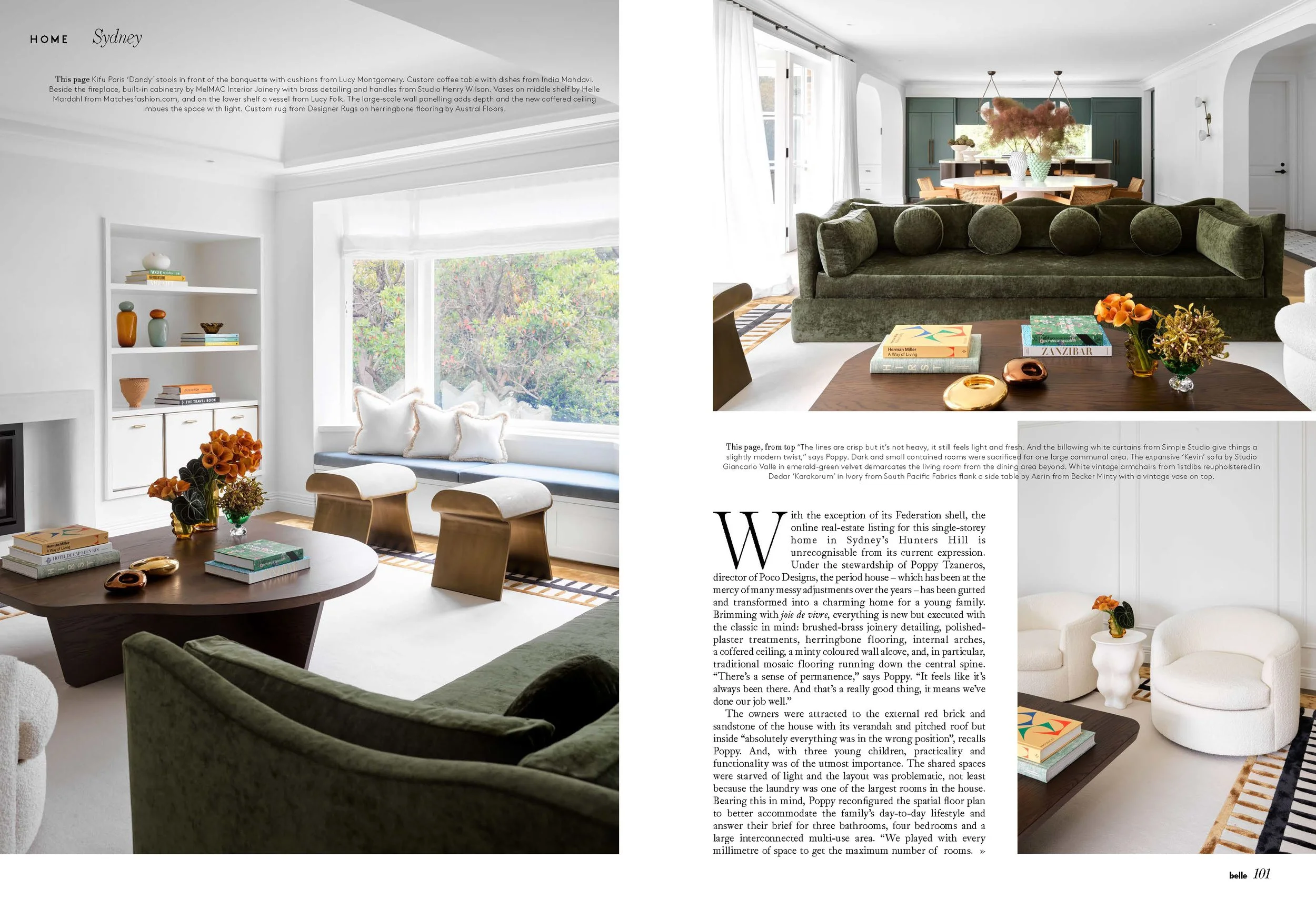 Interior view of a bright, modern living room with a round wooden coffee table holding books, vases, and flowers. There are two cream upholstered stools and a green velvet sofa with multiple large round cushions. Large windows with white curtains allow natural light to fill the space. The room features white walls, a built-in bookshelf with decorative items, and a high ceiling with simple crown molding.