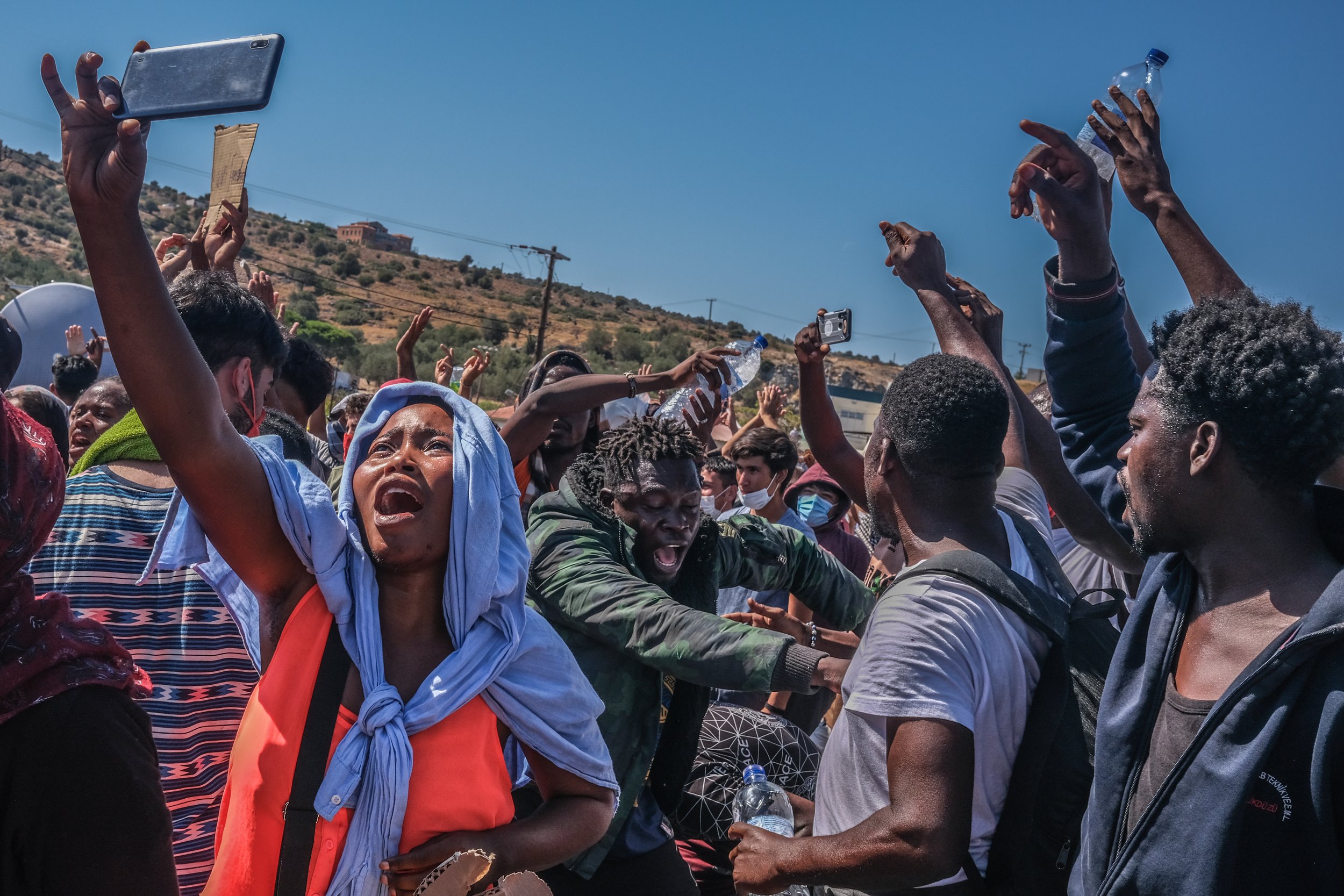 Crowd of people protesting or demonstrating outdoors under sunny sky, expressing anger and protest, some holding bottles and phones, with hills and power lines in background.