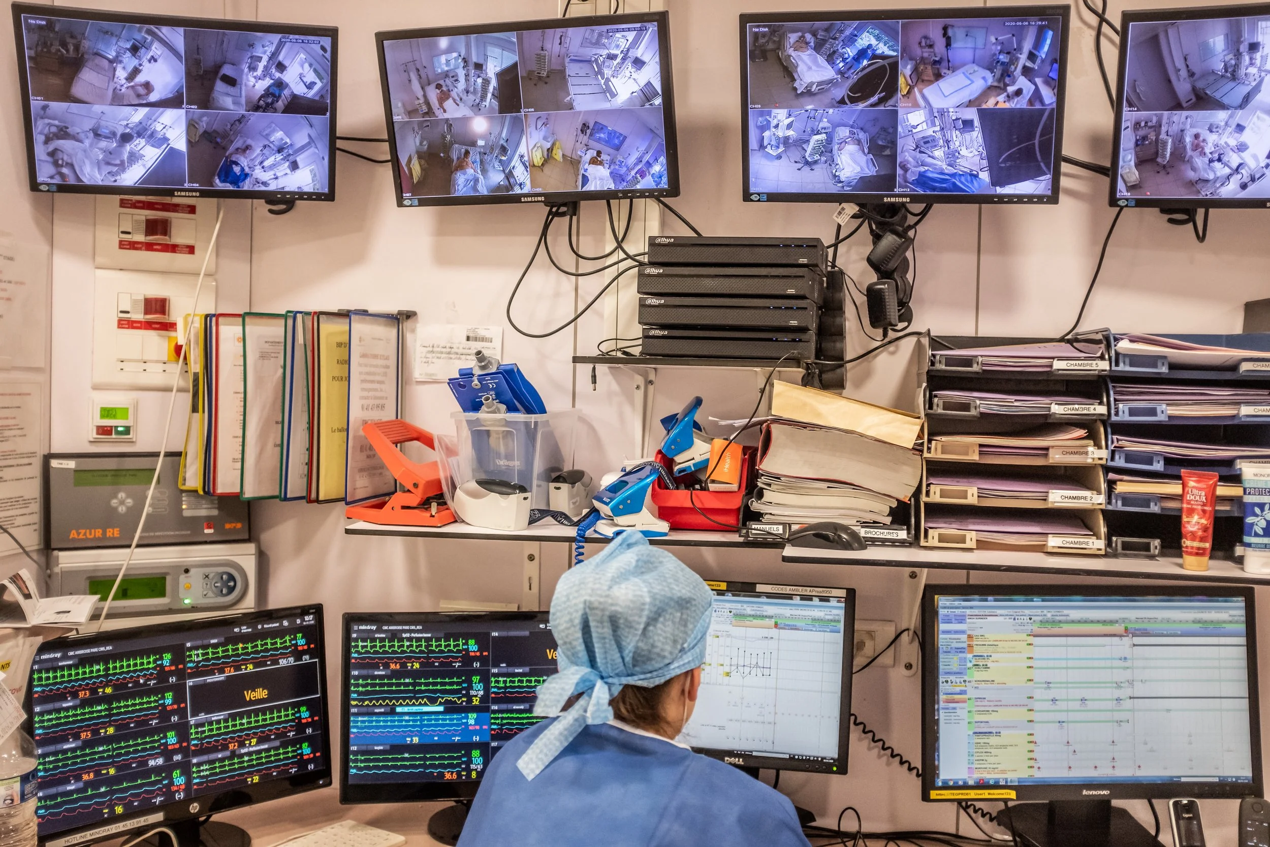 Hospital monitoring room with multiple screens displaying patient vitals and medical images, a healthcare worker wearing a surgical cap working at a computer.