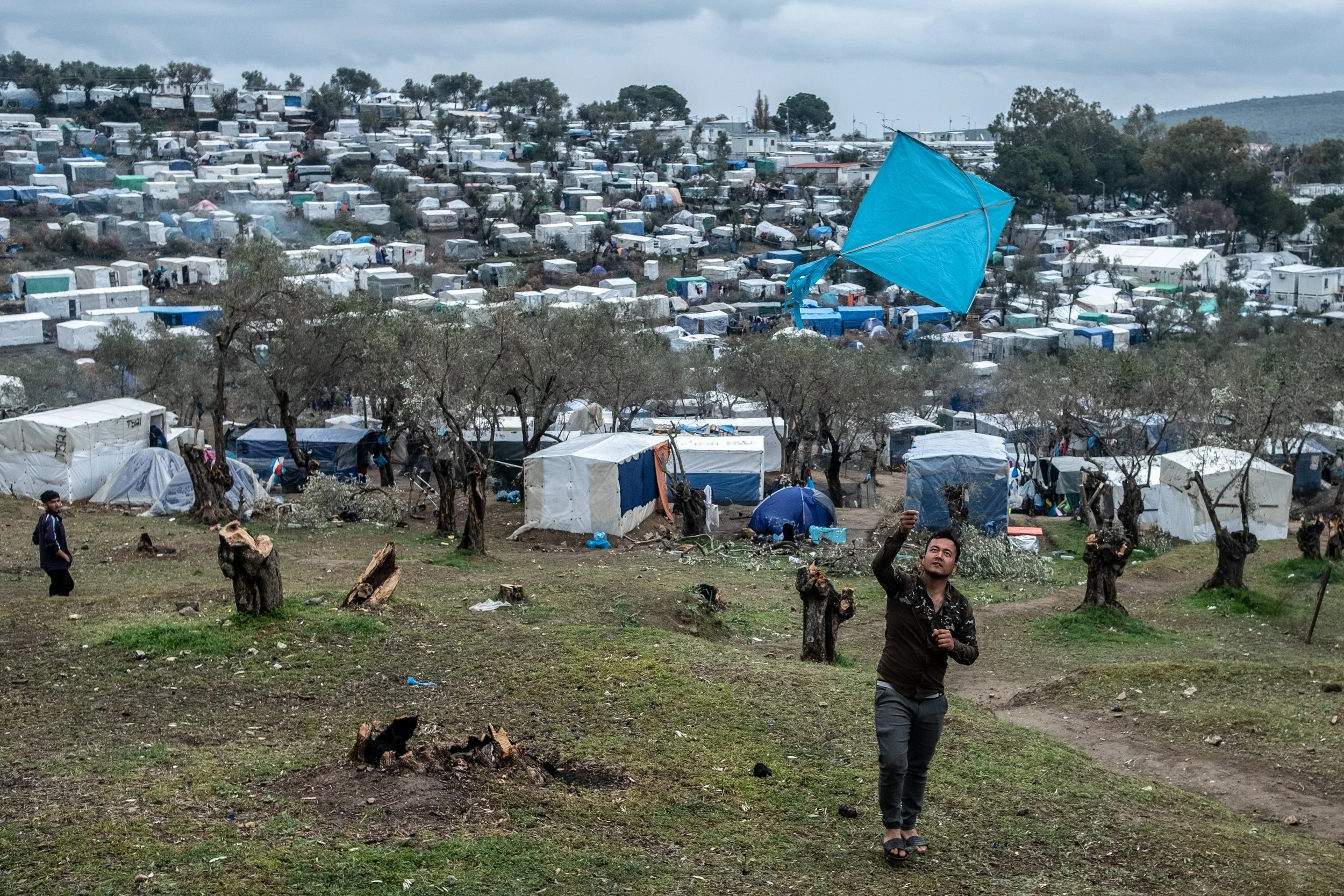 A man flying a kite in a refugee camp with numerous tents and makeshift shelters, some trees, and a cloudy sky.