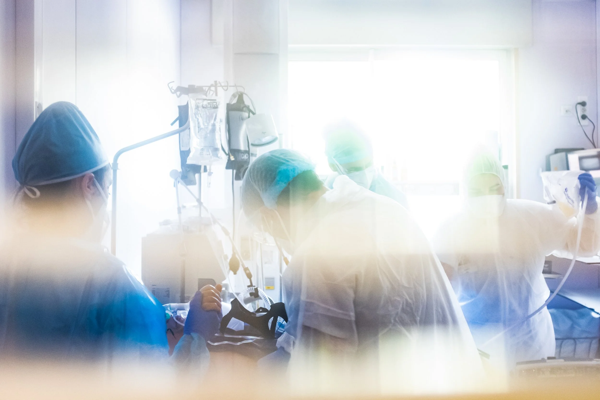 Medical professionals in scrubs and masks gathered around a patient in a hospital room, with medical equipment and sunlight coming through a window in the background.