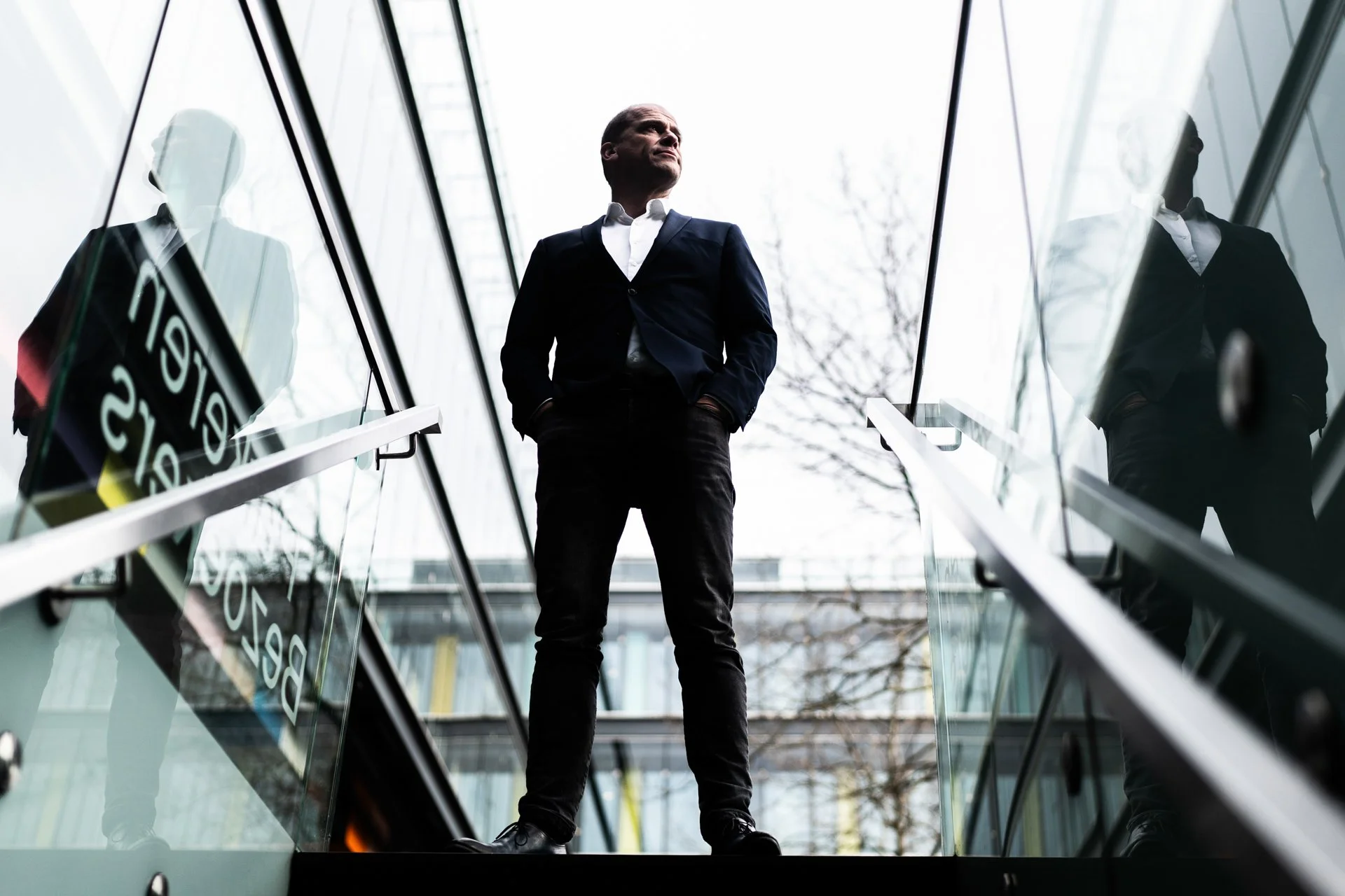 A man in a formal suit standing on an escalator, looking upward with hands in pockets, with glass walls reflecting him and a cityscape background.