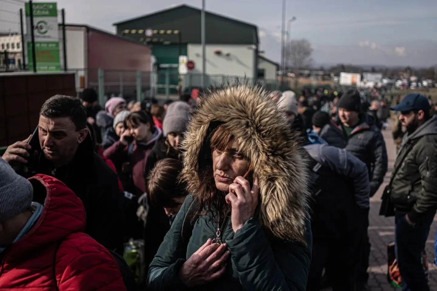 Group of people, some on phones, standing outside in a crowded area; one woman with a fur-lined hood in the center appears to be on the phone.