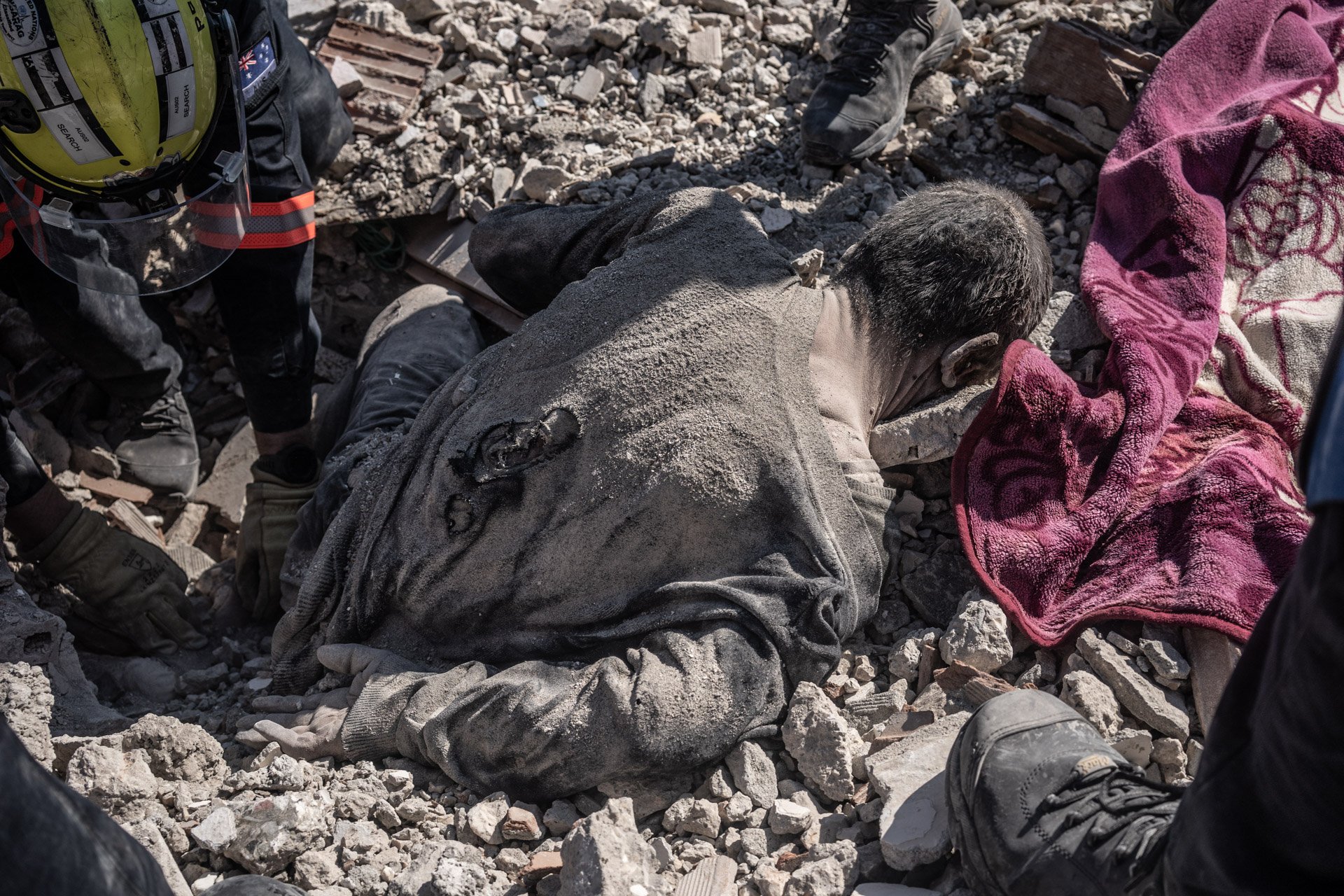 A man lying on the ground covered in dust and debris, surrounded by rubble, with rescue workers nearby.