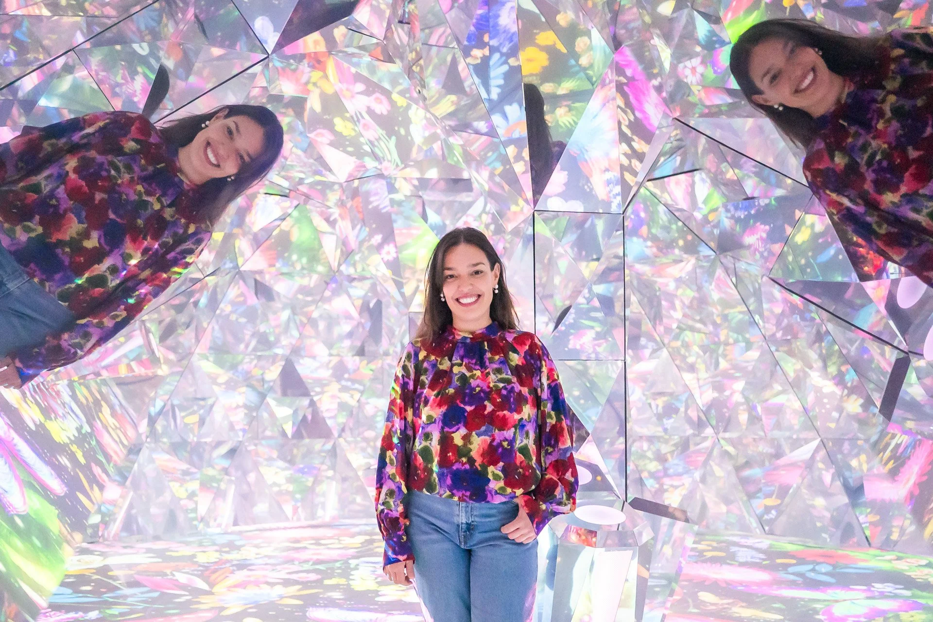 Woman smiling in an iridescent, multi-colored, geometric art installation with mirror reflections.