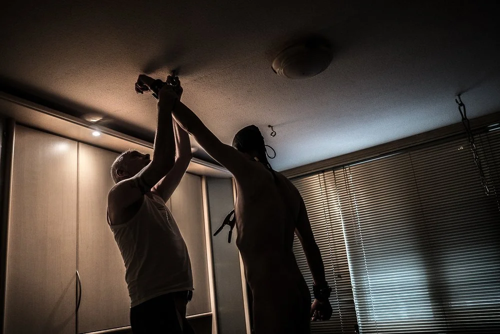 Two people silhouetted against a window with blinds, one helping the other to install or fix a ceiling light fixture.