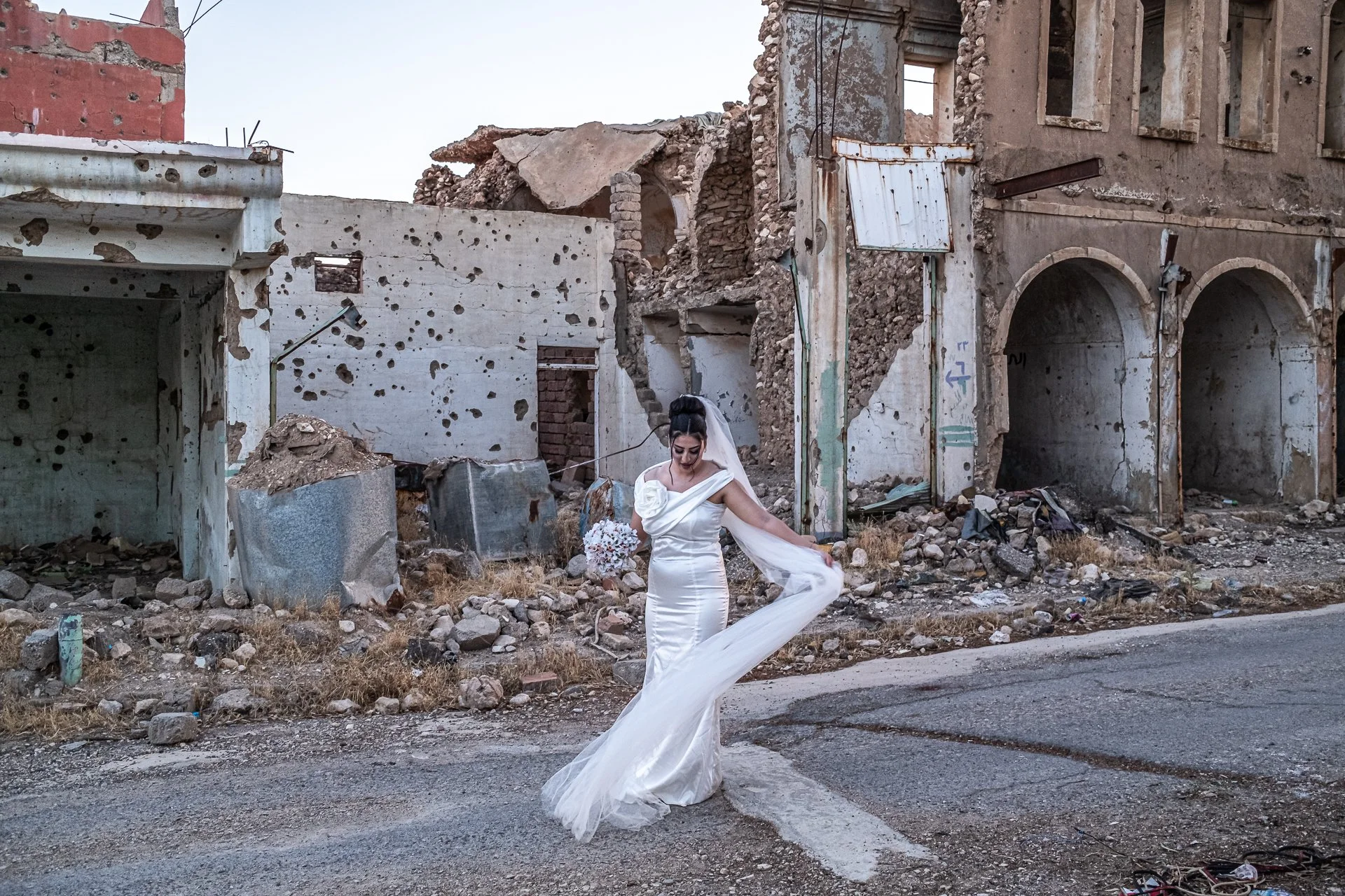 A woman in a white wedding dress and veil stands on an empty road, surrounded by destroyed and abandoned buildings with walls riddled with holes.