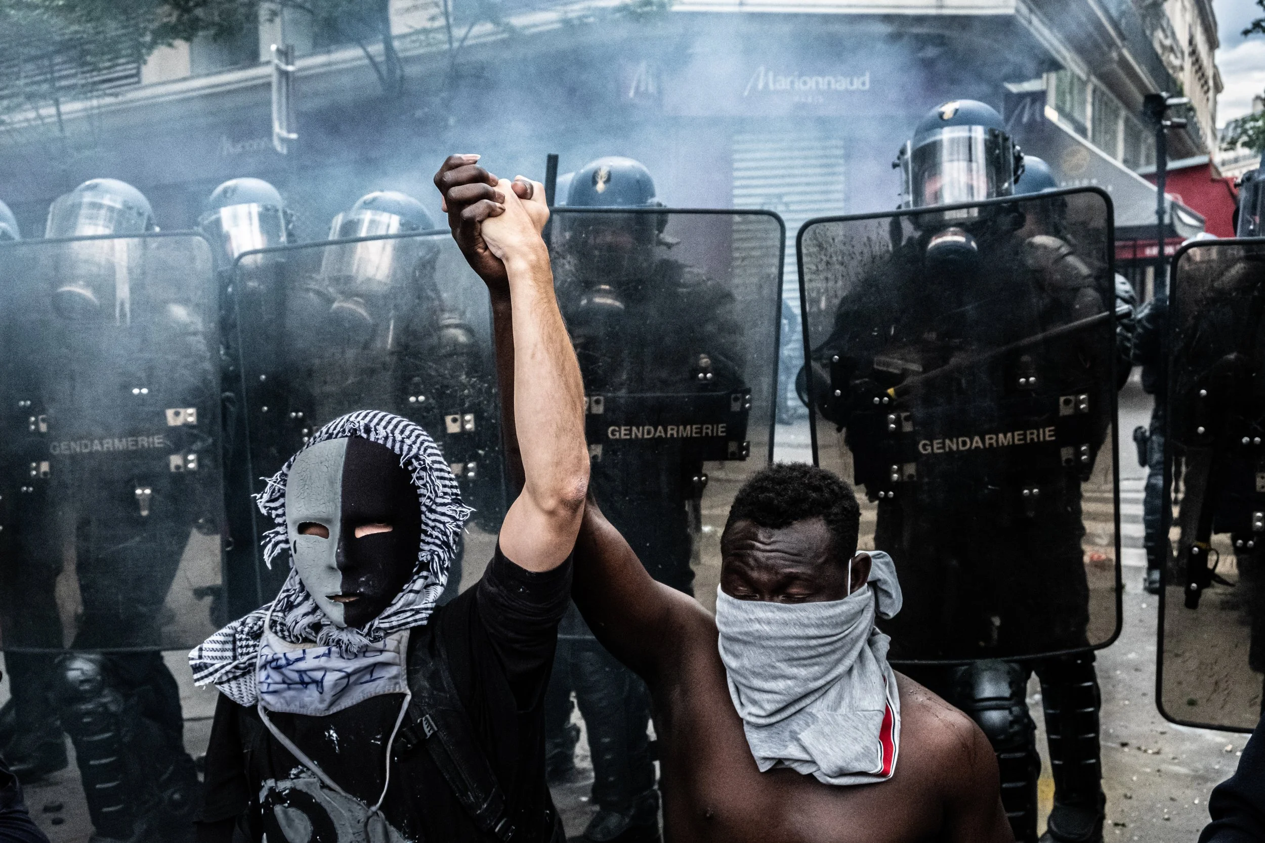 Two protesters with masked faces and bandanas around their necks are holding hands in front of a line of riot police with shields labeled 'Gendarmerie.' Smoke or tear gas is in the air.
