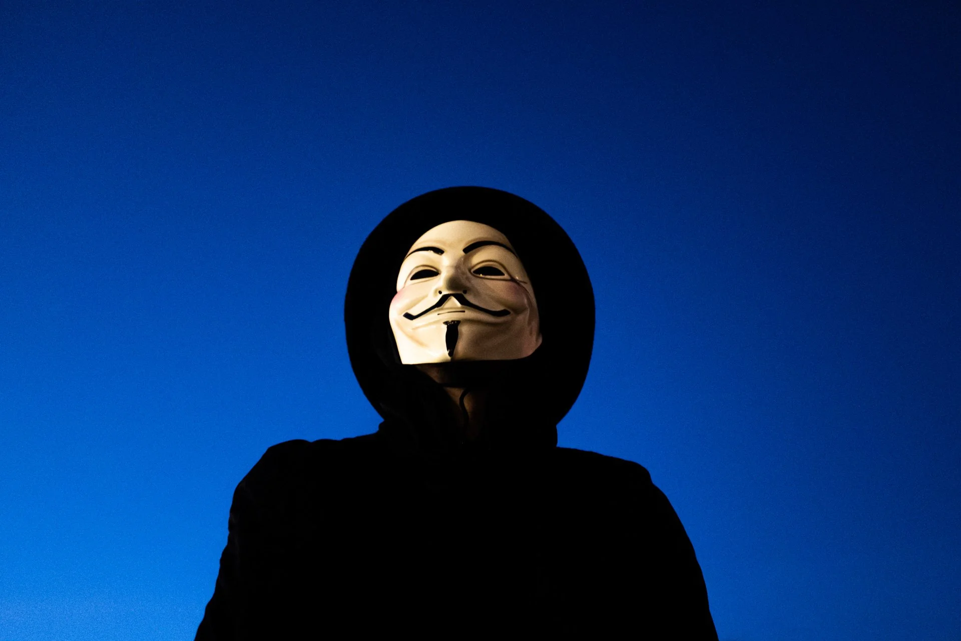 Person wearing a Guy Fawkes mask and black hoodie against a deep blue sky at night.