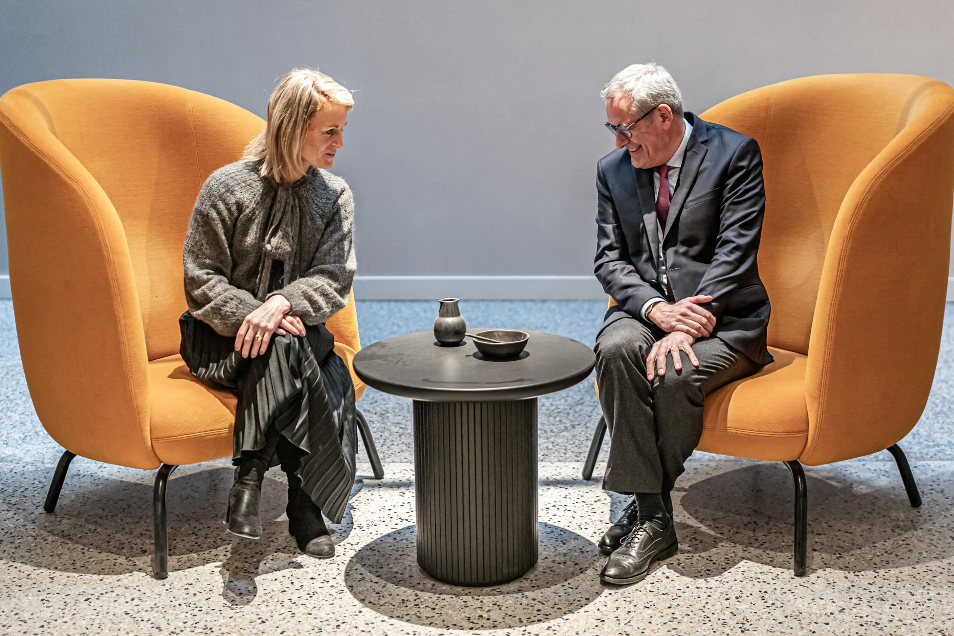 Two people sitting in orange high-backed chairs facing each other across a small black round table with a small vase and a bowl, engaged in conversation, indoors with blue carpet and plain wall background.