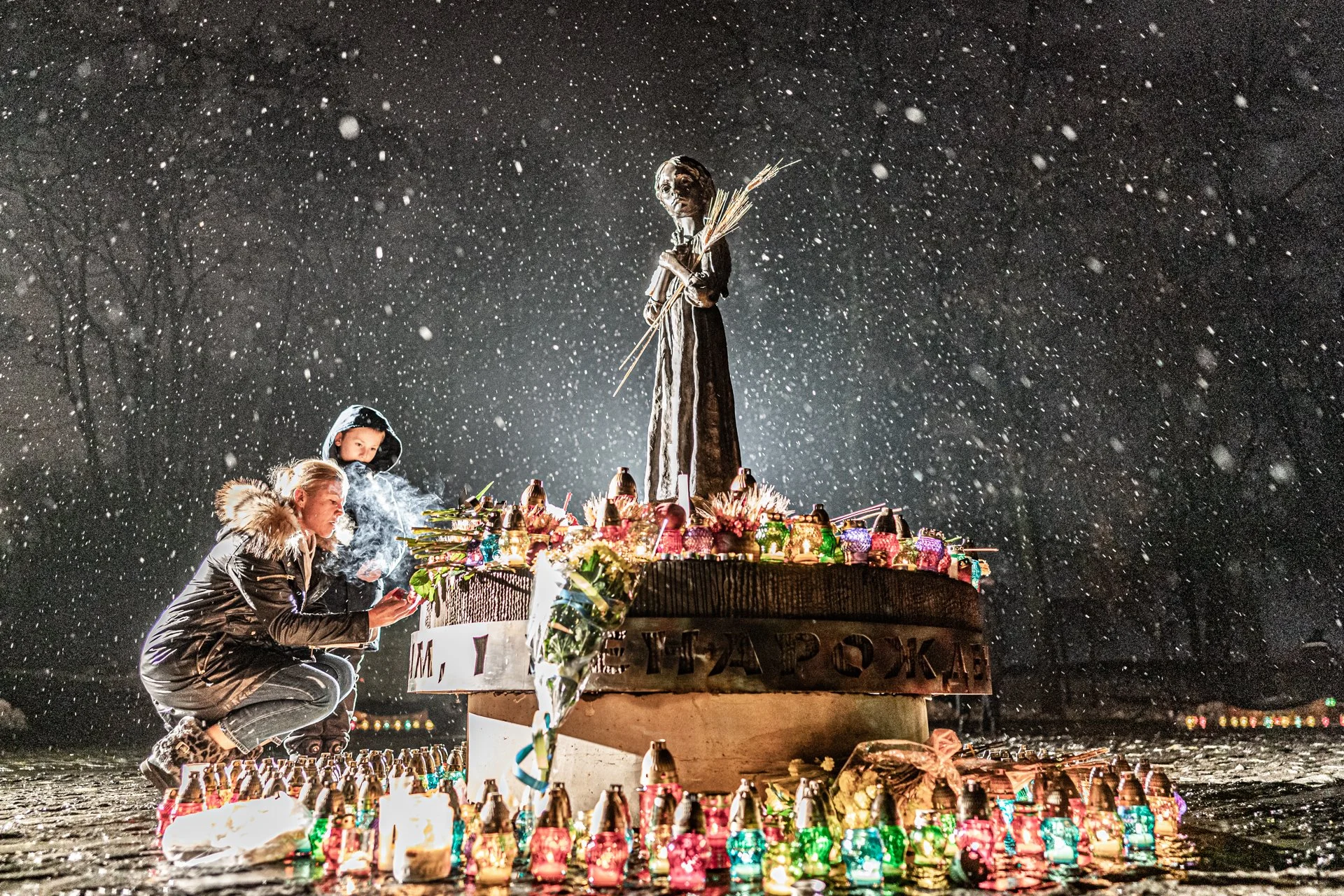 People celebrating at a nighttime event with a large statue on a decorated pedestal surrounded by colorful lamps and candles, snow falling, two women and a child near the statue.