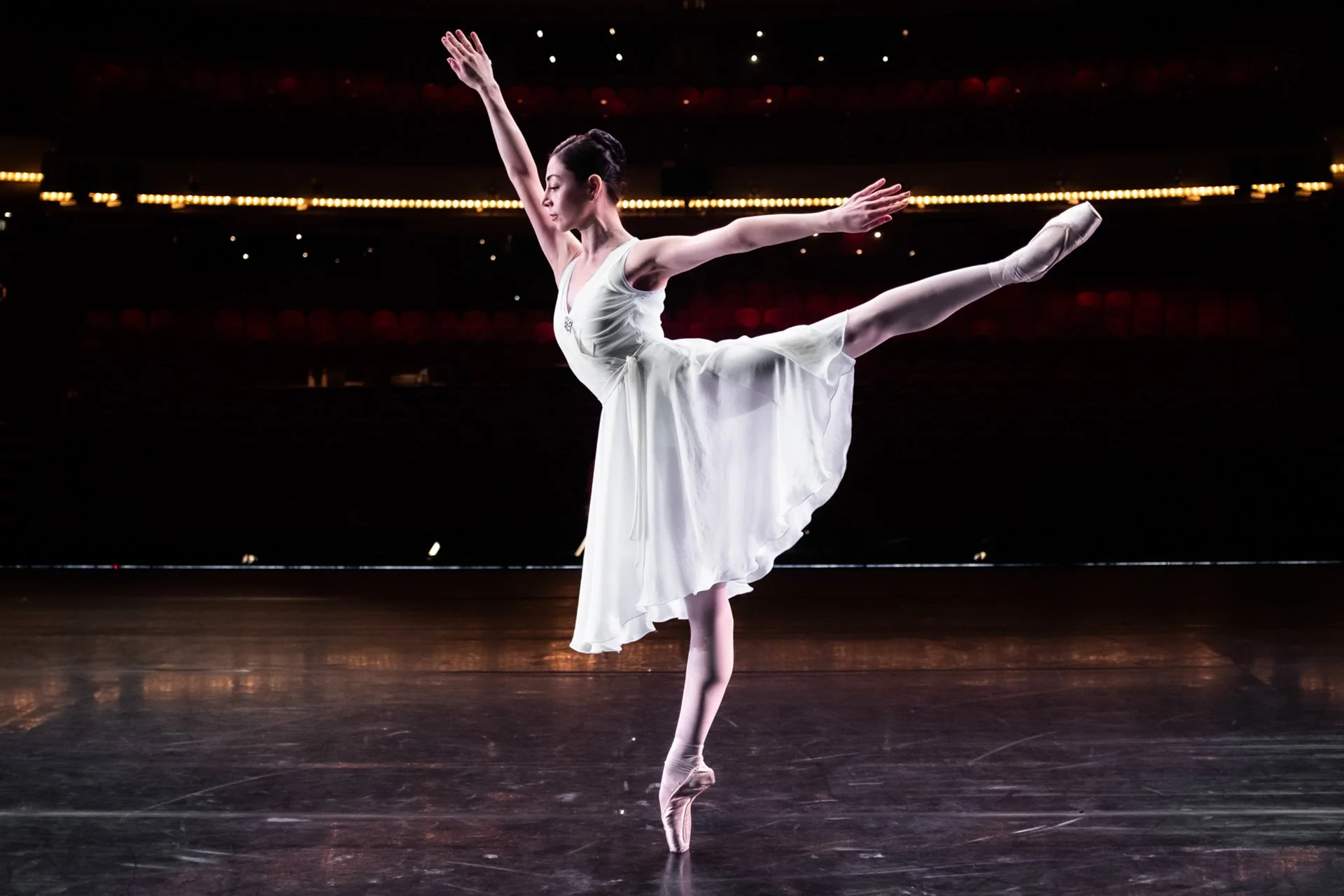 Ballet dancer in a white dress performing an arabesque on stage with a dark background.