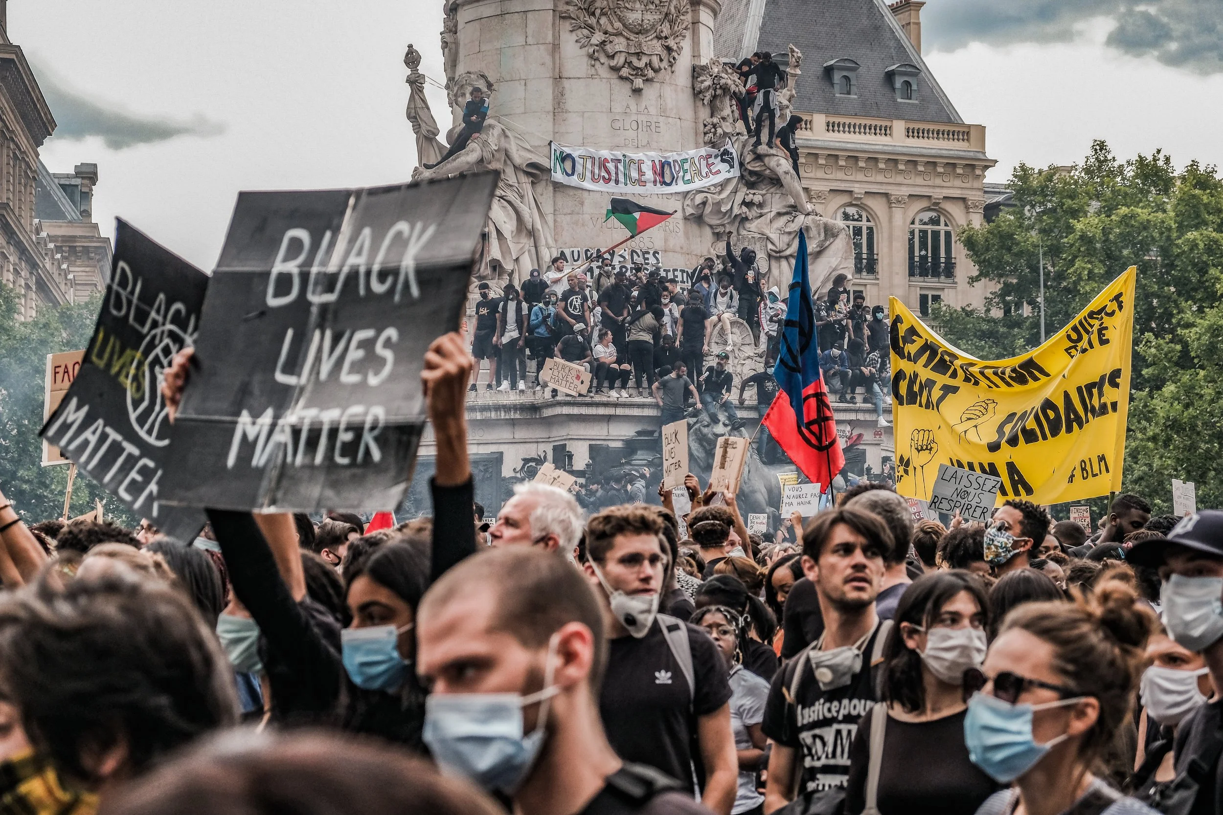 A large crowd of protesters gathered at a demonstration, holding up signs and banners with messages such as "Black Lives Matter" and advocating for justice and peace. The Eiffel Tower's replica and a monument with sculptures are visible in the backgr