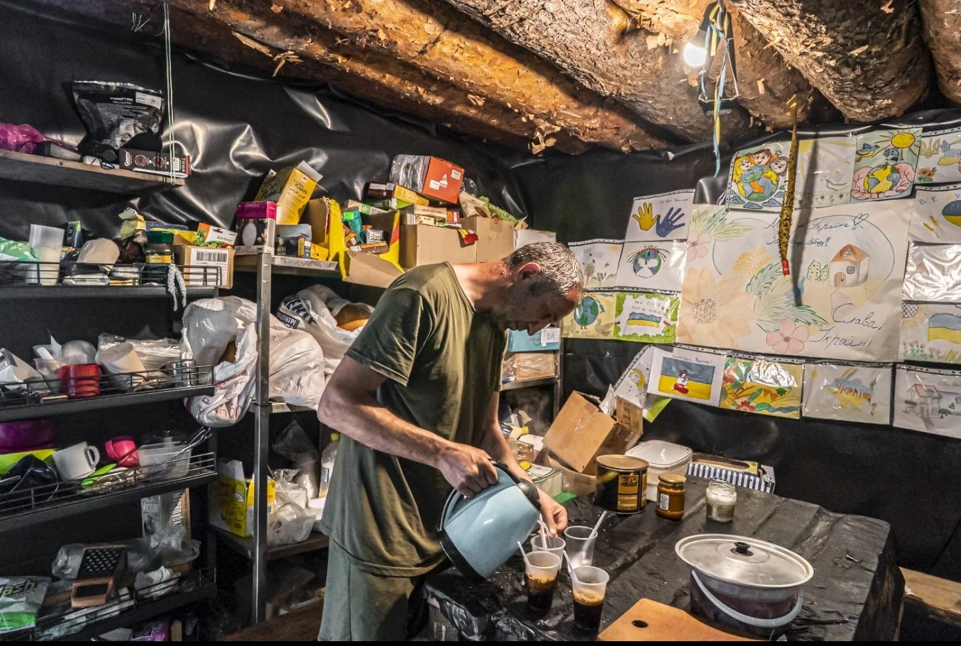 A man pouring drinks into glasses in a room with shelves of supplies and colorful drawings on the wall.