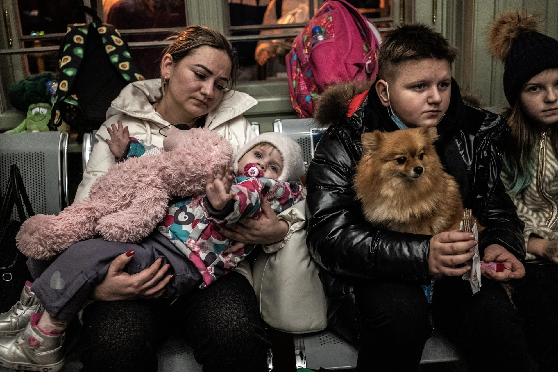 A woman sitting with a young girl on her lap, holding a teddy bear, in a public transportation setting with other children and backpacks nearby.
