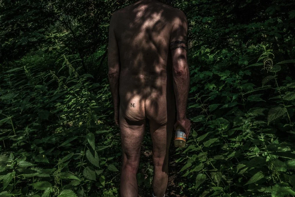 Naked man walking away in a lush green forest, holding a bottle in his right hand, with a tattoo on his lower back that says 'NE'.