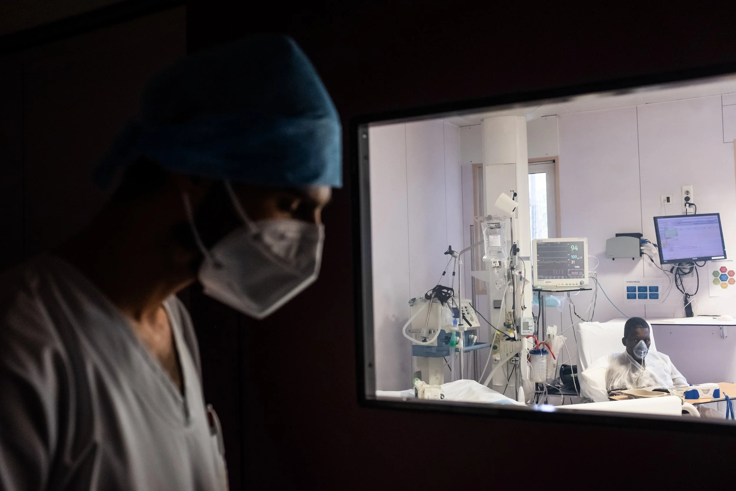A healthcare worker wearing a disposable mask and blue surgical cap looks through a window into a hospital room where a patient is lying in bed, connected to medical equipment.