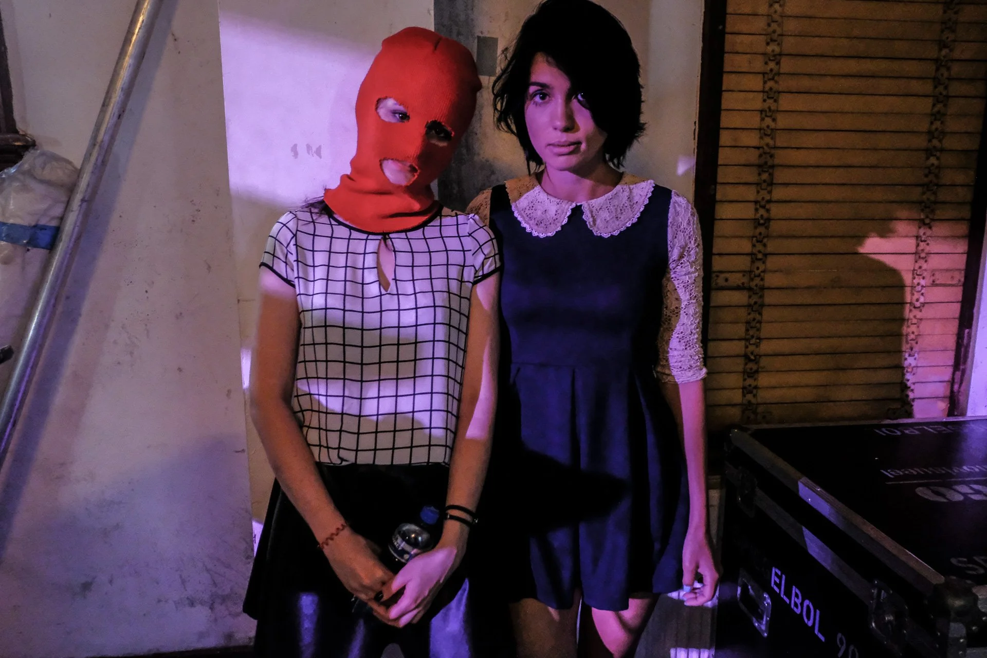 Two women standing close together indoors, one wearing a red ski mask and a checkered top, holding a water bottle, the other with short dark hair in a black lace dress with a white collar, in a dimly lit setting with colored lighting.