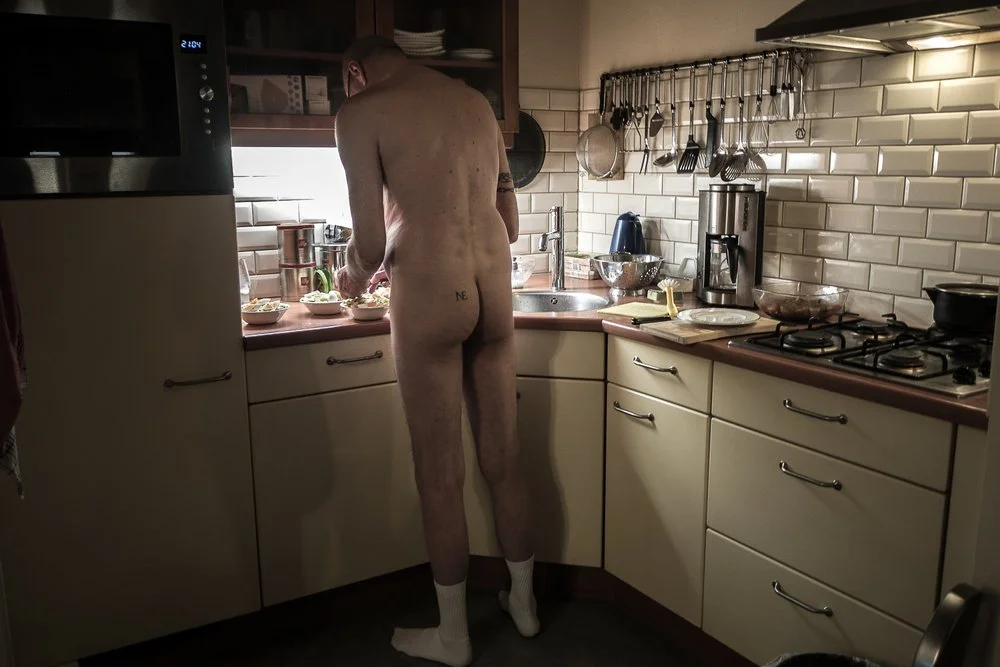 A naked man wearing socks, with a tattoo, standing in a kitchen near the counter, preparing food. The kitchen has white cabinets, a tiled backsplash, and various utensils and appliances.