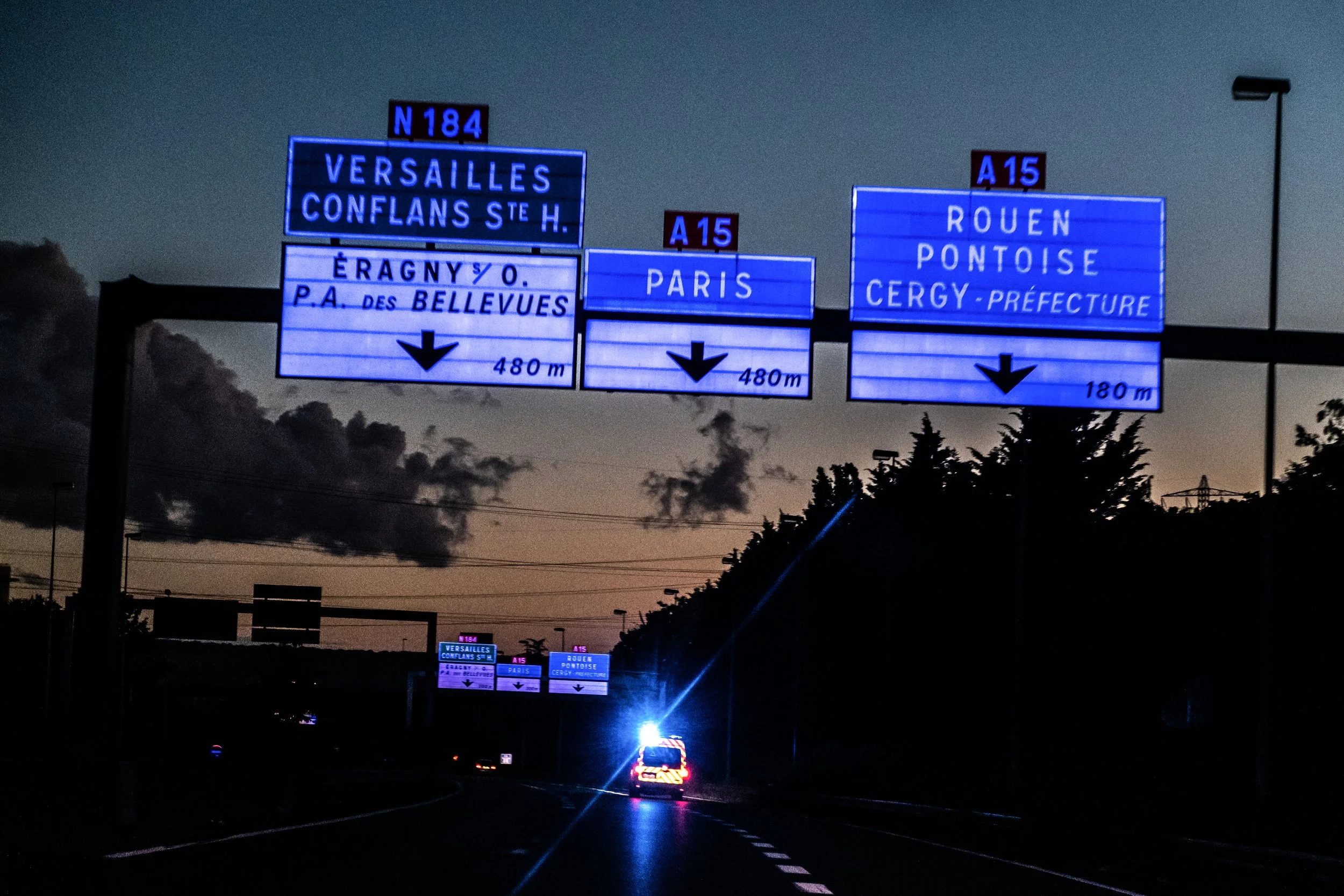 Nighttime highway with illuminated blue overhead road signs indicating directions to Versailles, Paris, Rouen, Pontoise, and Cergy-Prefecture, with distance markers of 480 meters and 180 meters.