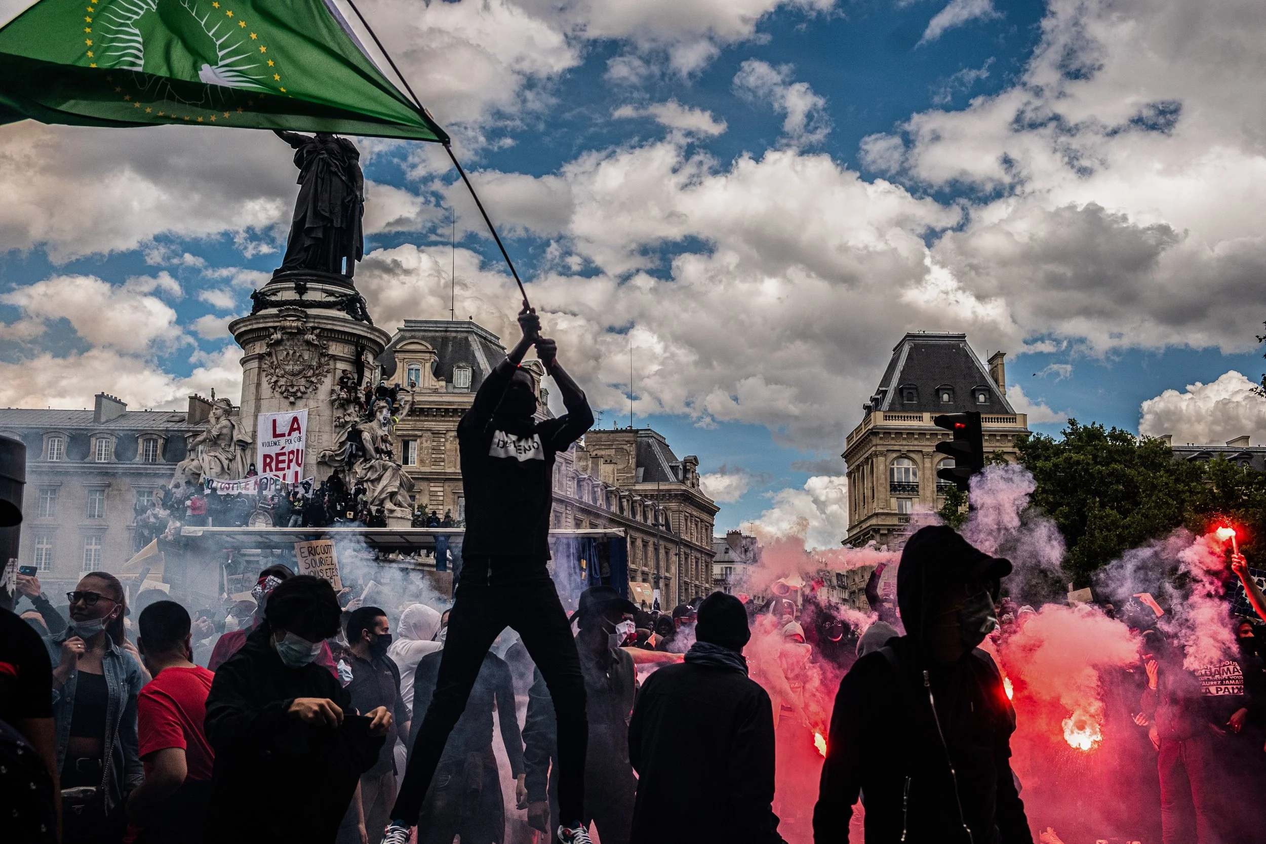 Crowd of protesters on a city street holding signs, with some using smoke bombs and flares, in front of historic buildings and a statue under a cloudy sky.