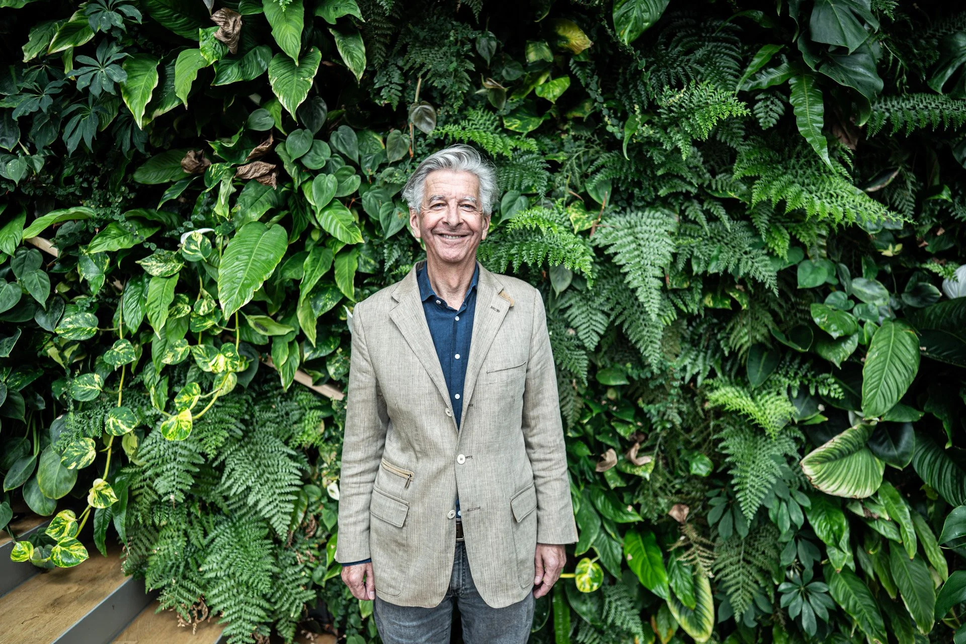 An older man with gray hair and a smiling expression, dressed in a beige blazer over a dark blue shirt, standing in front of a lush green wall of various tropical plants and ferns.