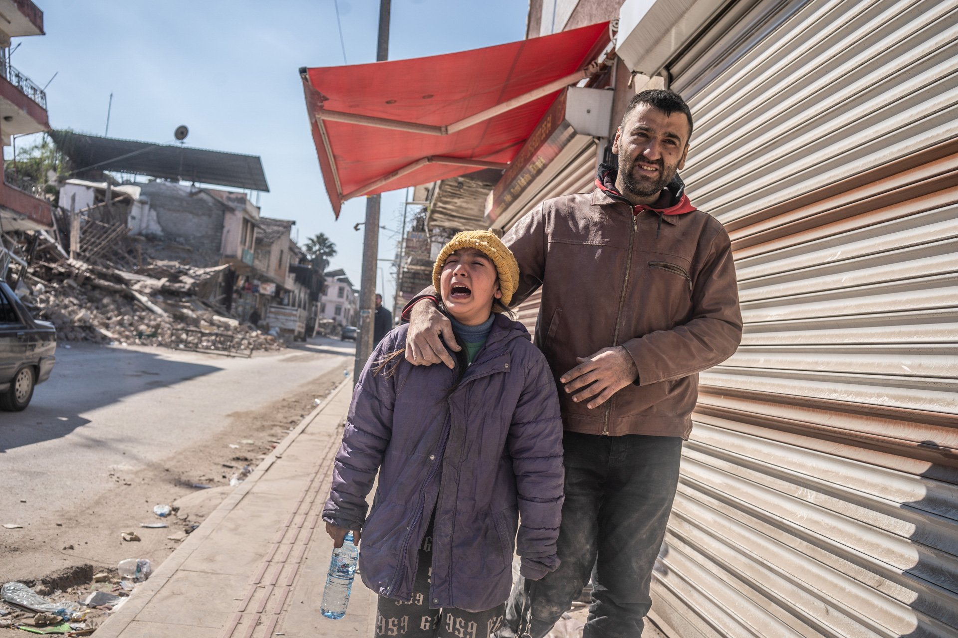 A man and a young girl are standing on a sidewalk, with the girl crying and the man comforting her. The background shows damaged buildings and debris, indicating a disaster or conflict area.