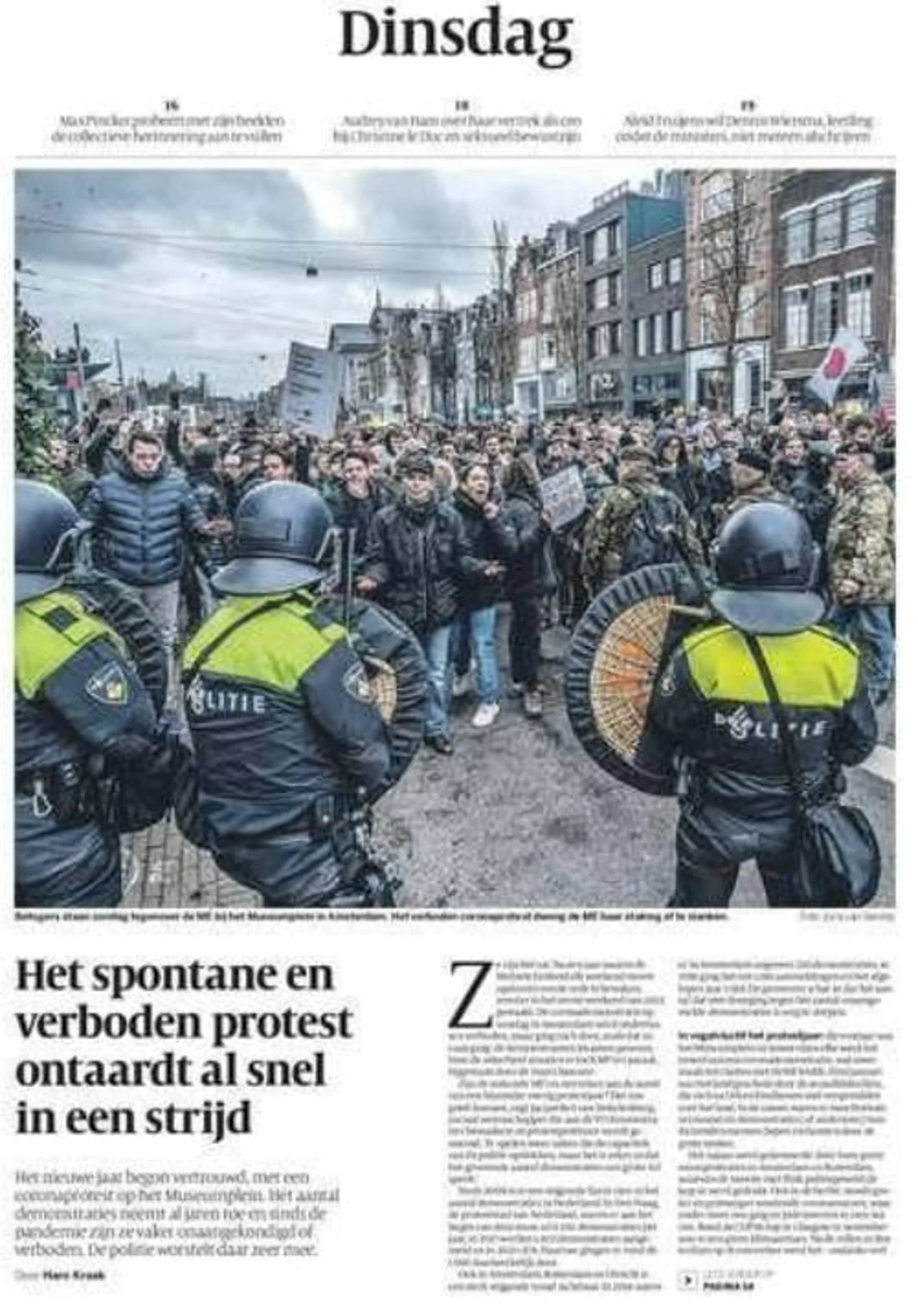 A large protest with people holding signs and flags in a street, facing police officers in riot gear with shields, in an urban area with buildings and cloudy sky.