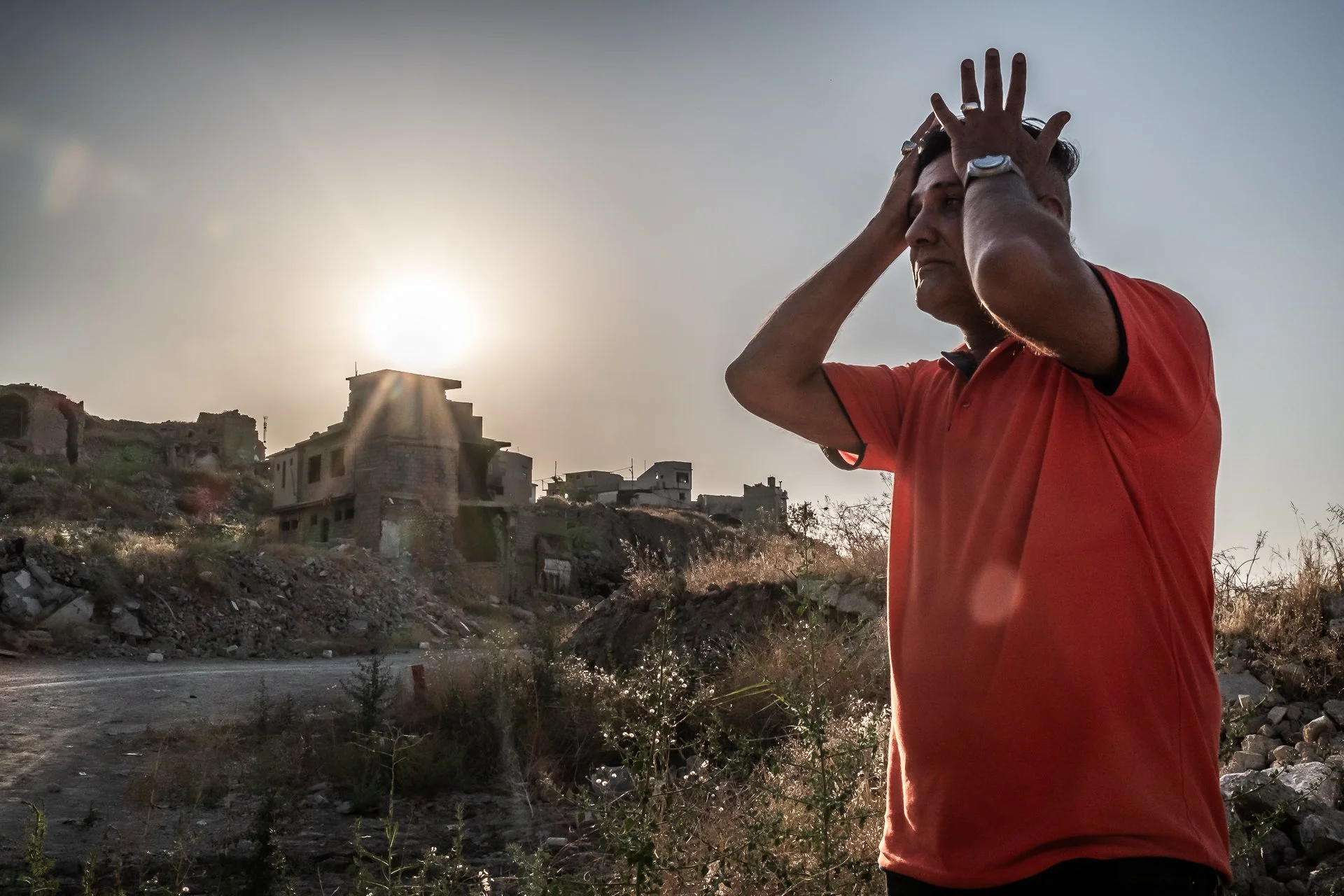 A man in a red shirt stands outdoors at sunset with hands on his head, looking contemplative, with abandoned buildings on a hillside in the background.
