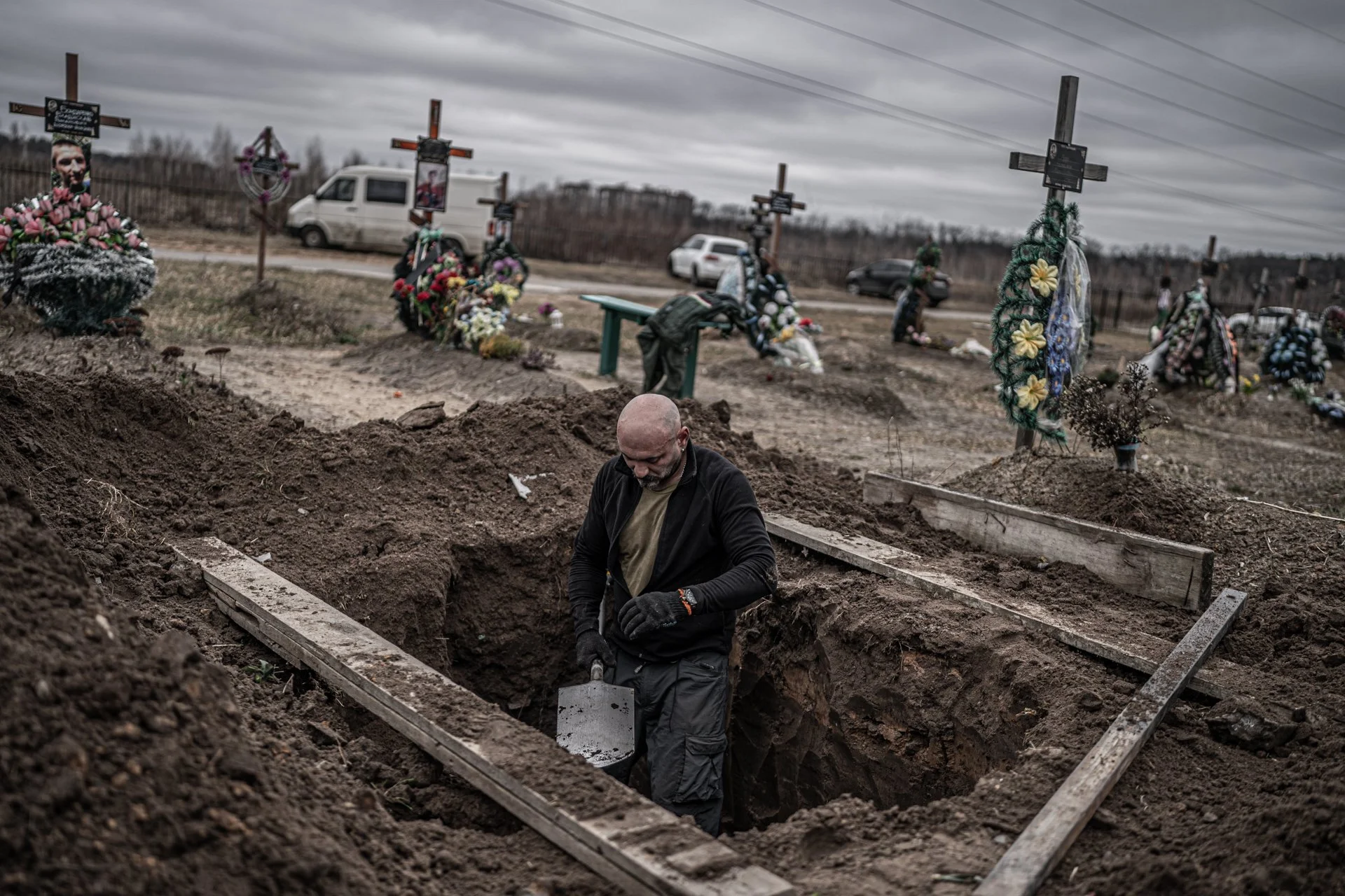 A man with a bald head and beard digging a grave in a cemetery during overcast weather. Graves are marked with crosses and decorated with flowers, and there are cars and a wooden fence in the background.
