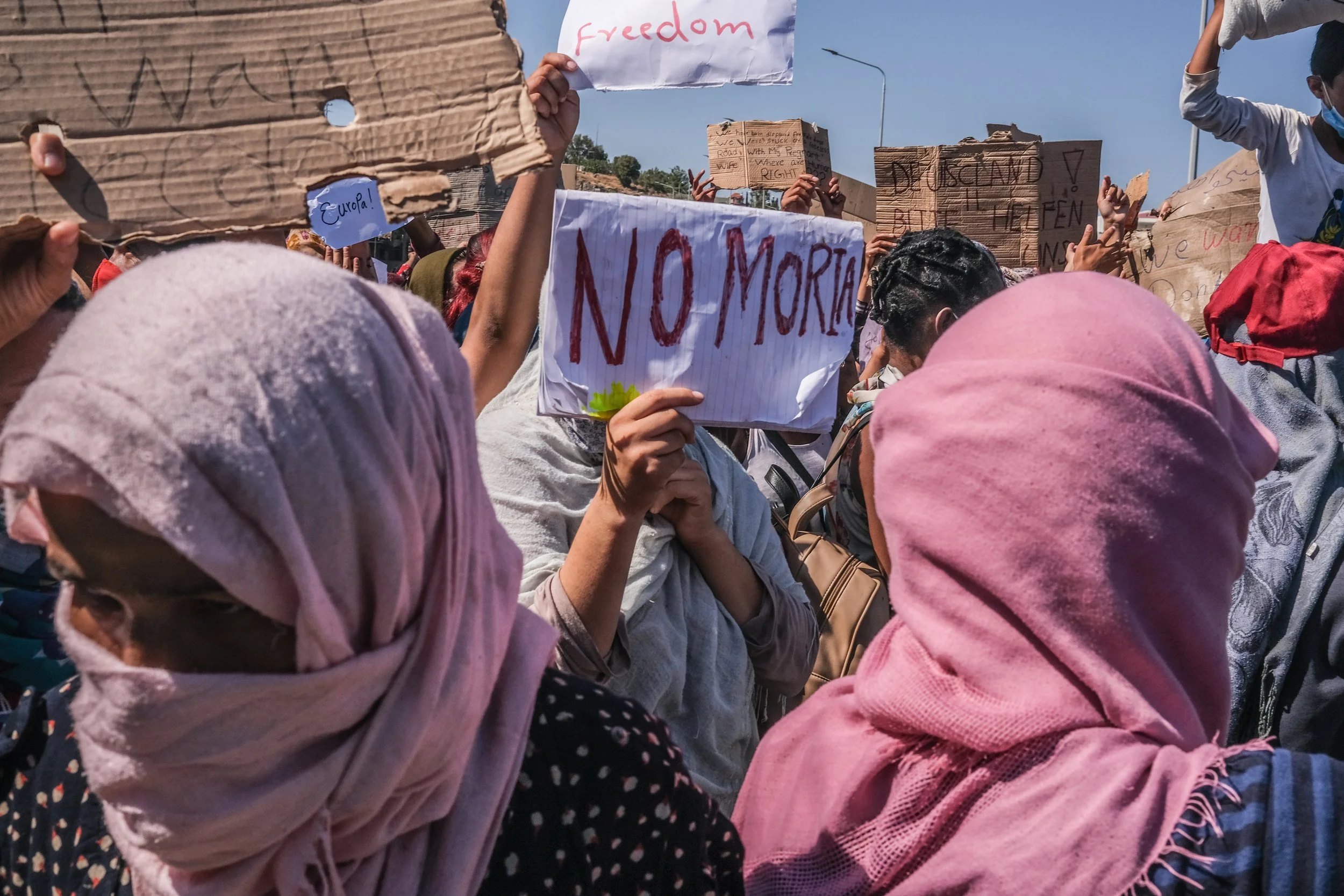 People participating in a protest, many wearing headscarves, holding signs with messages such as 'NO MORA' and 'freedom' in a crowd.