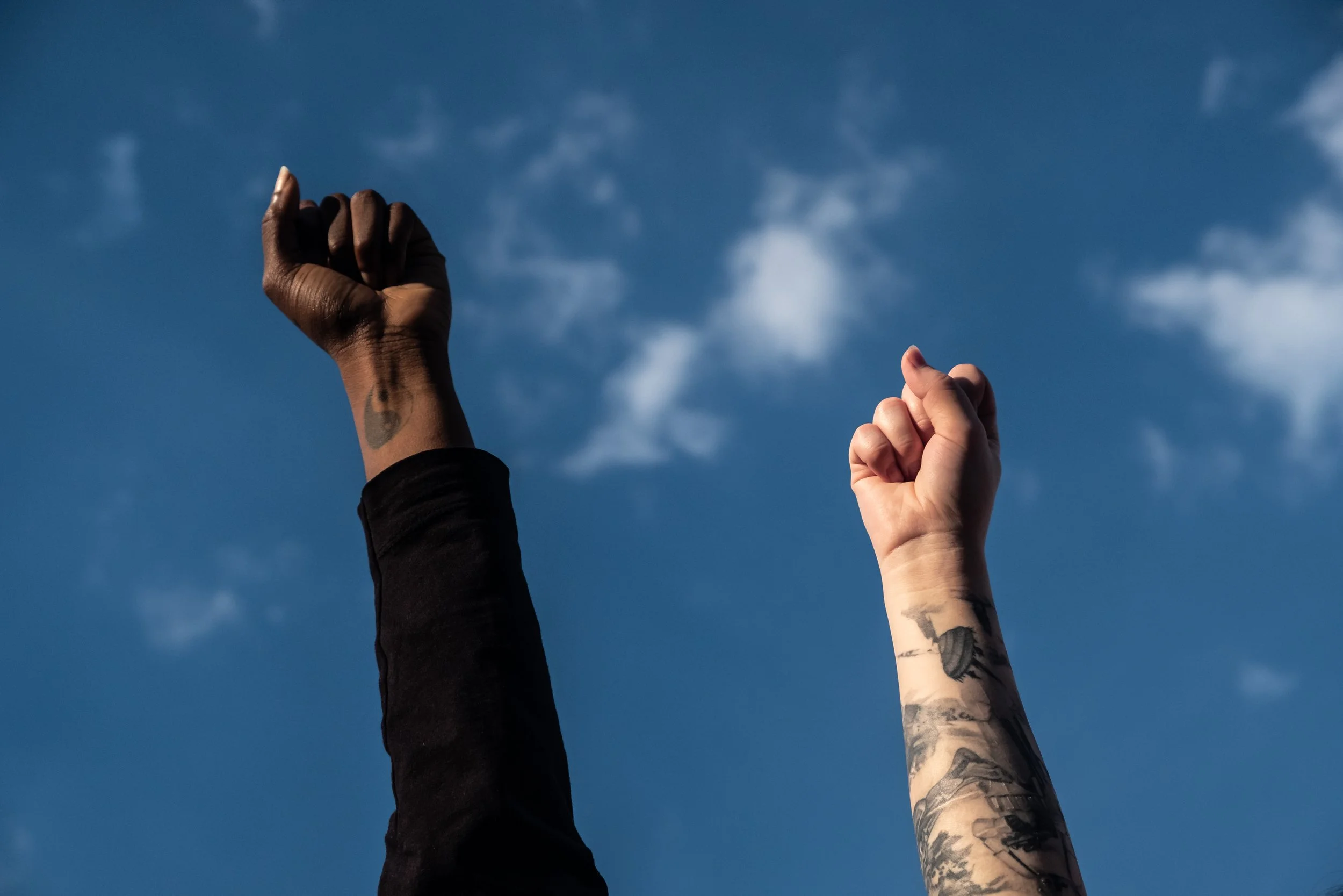 Two raised fists, one black and one with tattoos, against a blue sky with scattered clouds.